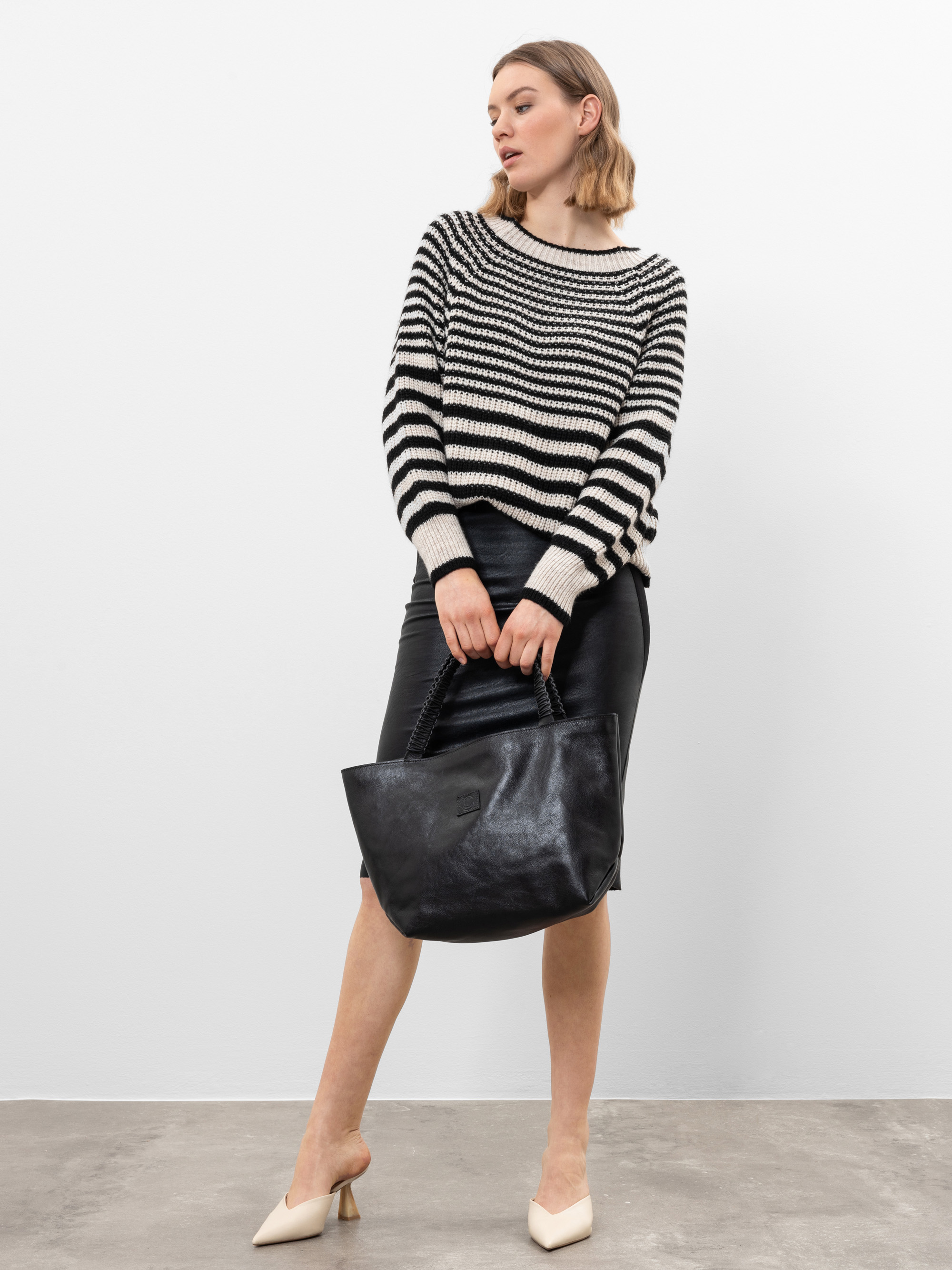 Eine Frau in einem schwarz-weiß gestreiften Pullover, einem schwarzen Lederrock und weißen Absätzen steht mit einer großen schwarzen Handtasche vor einem schlichten weißen Wandhintergrund.