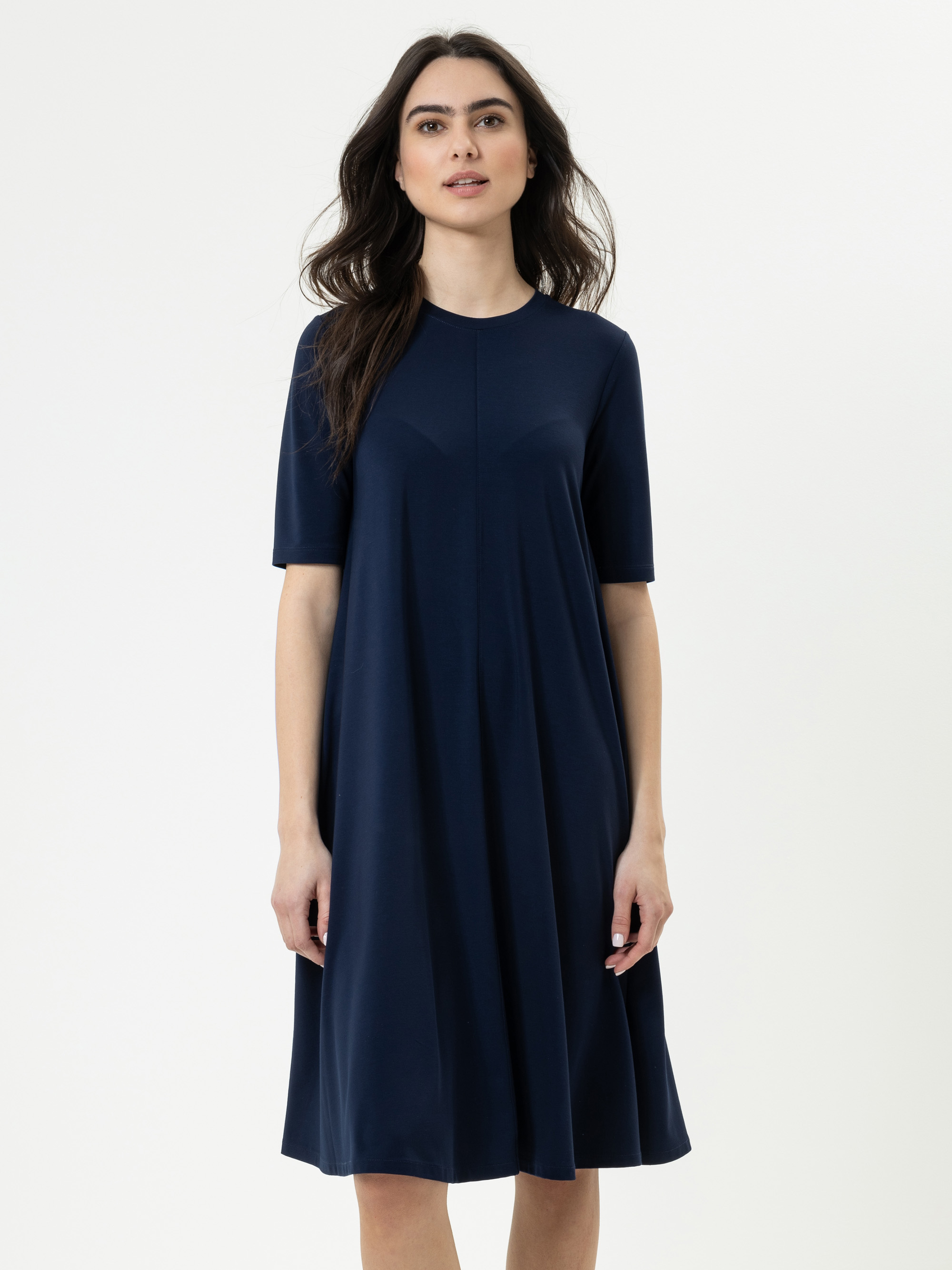 Eine Frau mit langen dunklen Haaren trägt ein marineblaues, knielanges, kurzärmeliges Kleid. Sie steht vor einem schlichten weißen Hintergrund und blickt mit neutraler Miene nach vorne.