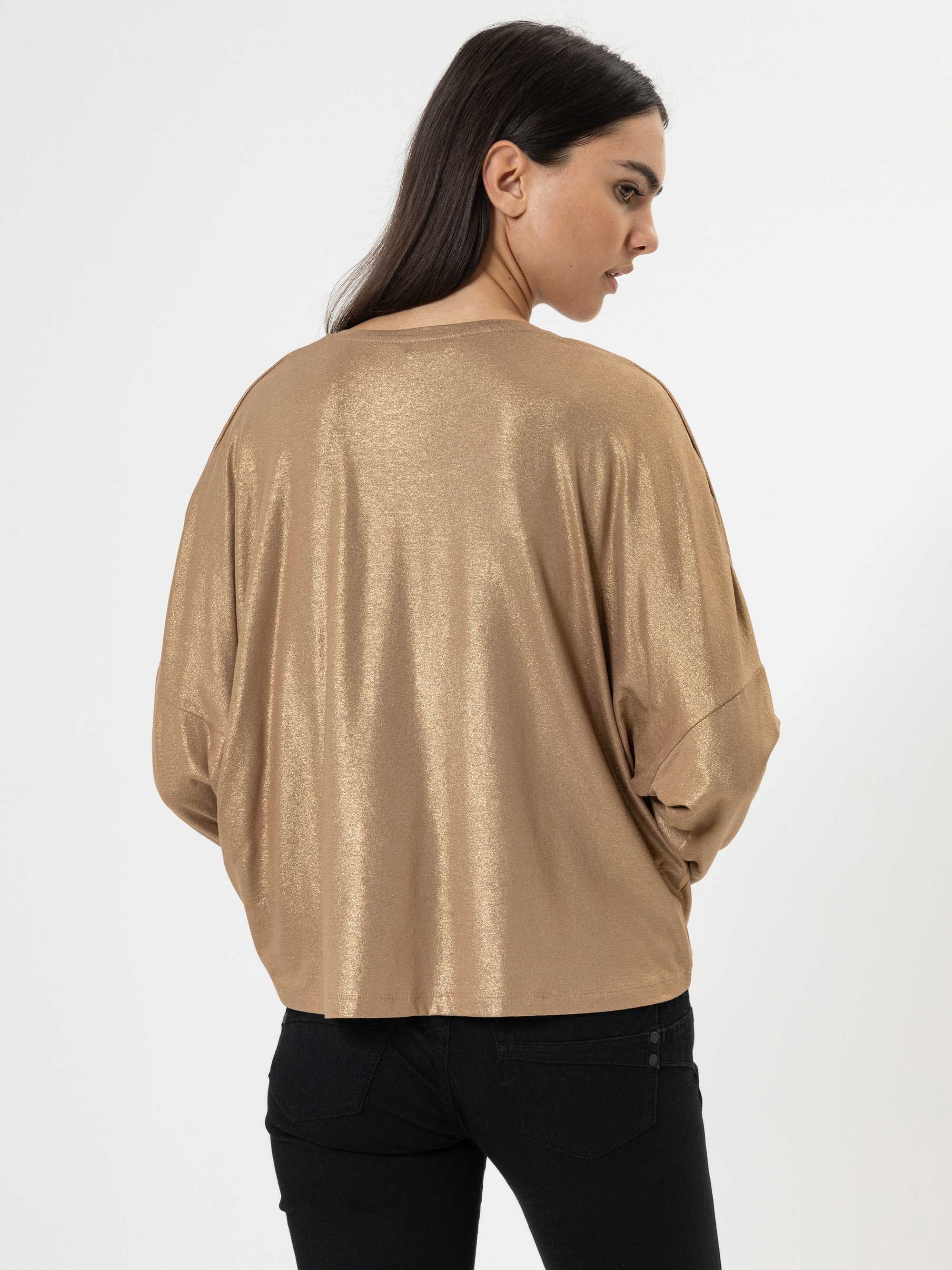 Eine Frau mit langen braunen Haaren, von hinten gesehen, trägt ein locker sitzendes, goldglänzendes Oberteil und schwarze Jeans vor einem schlichten weißen Hintergrund.