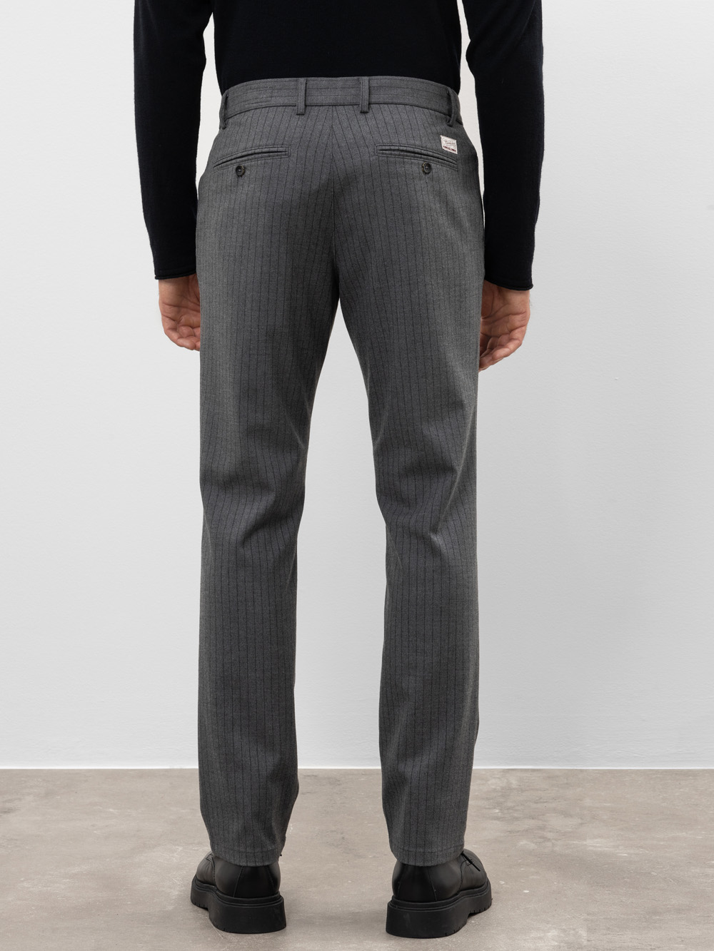 Eine Person, die eine graue Nadelstreifenhose, einen schwarzen Pullover und schwarze Schuhe trägt, steht auf einem hellgrauen Boden und blickt von der Kamera weg auf eine schlichte weiße Wand.