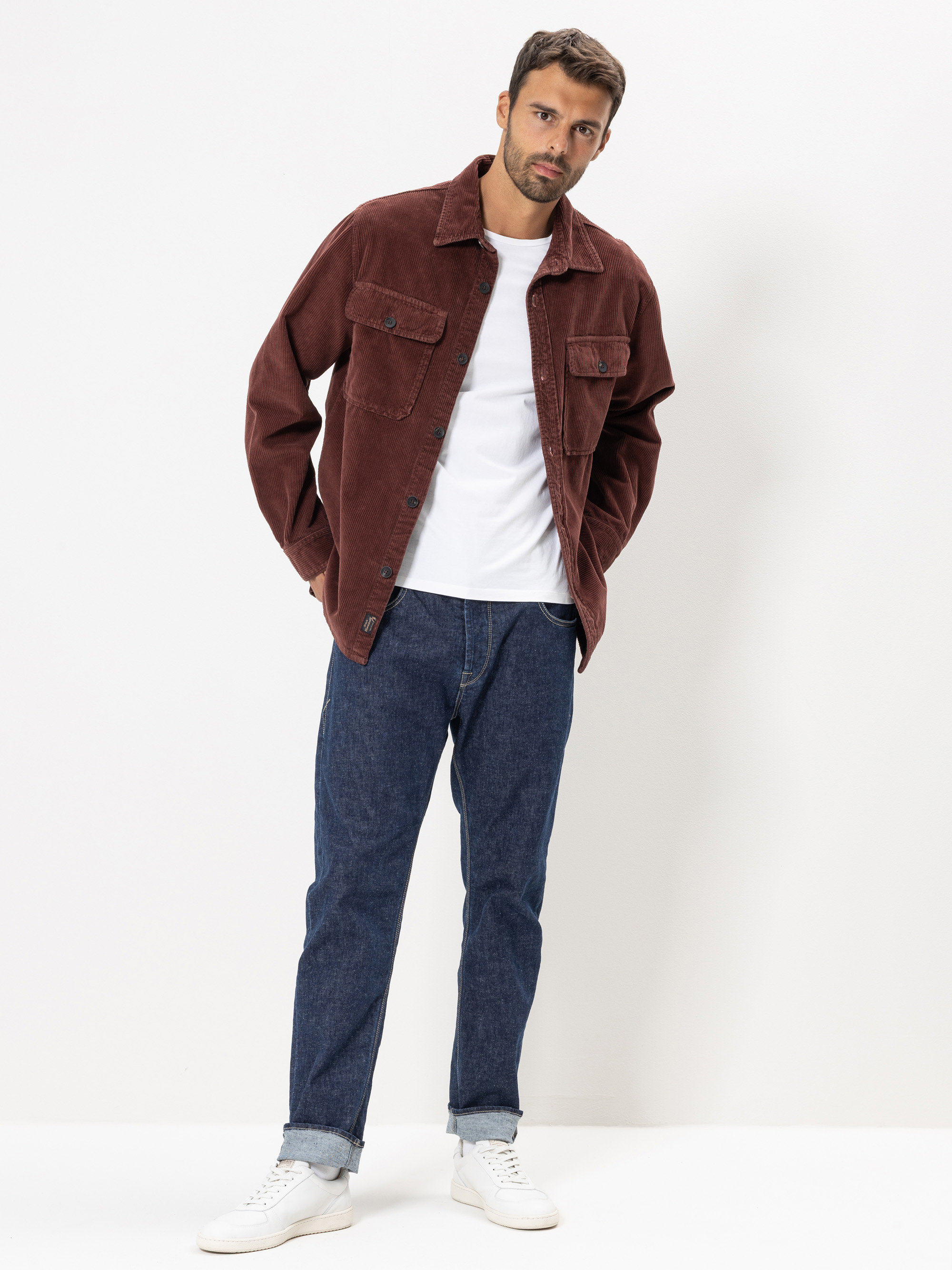 Ein Mann steht vor einem schlichten weißen Hintergrund. Er trägt eine kastanienbraune Cordjacke über einem weißen T-Shirt, blaue Jeans mit aufgerollten Bündchen und weiße Turnschuhe. Er hat die Hände in den Jackentaschen und ist leicht nach vorne g