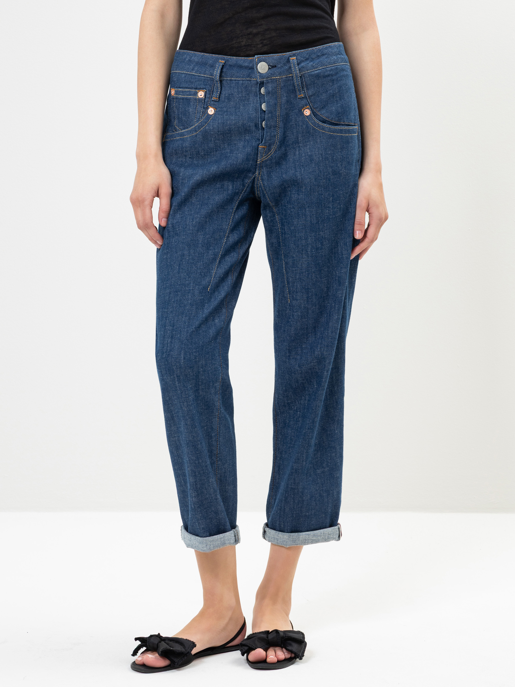 Eine Person, die hochgekrempelte blaue Jeans, ein schwarzes Oberteil und schwarze Schleifensandalen trägt, steht vor einem schlichten weißen Hintergrund. Das Bild zeigt das Outfit von den Schultern abwärts.