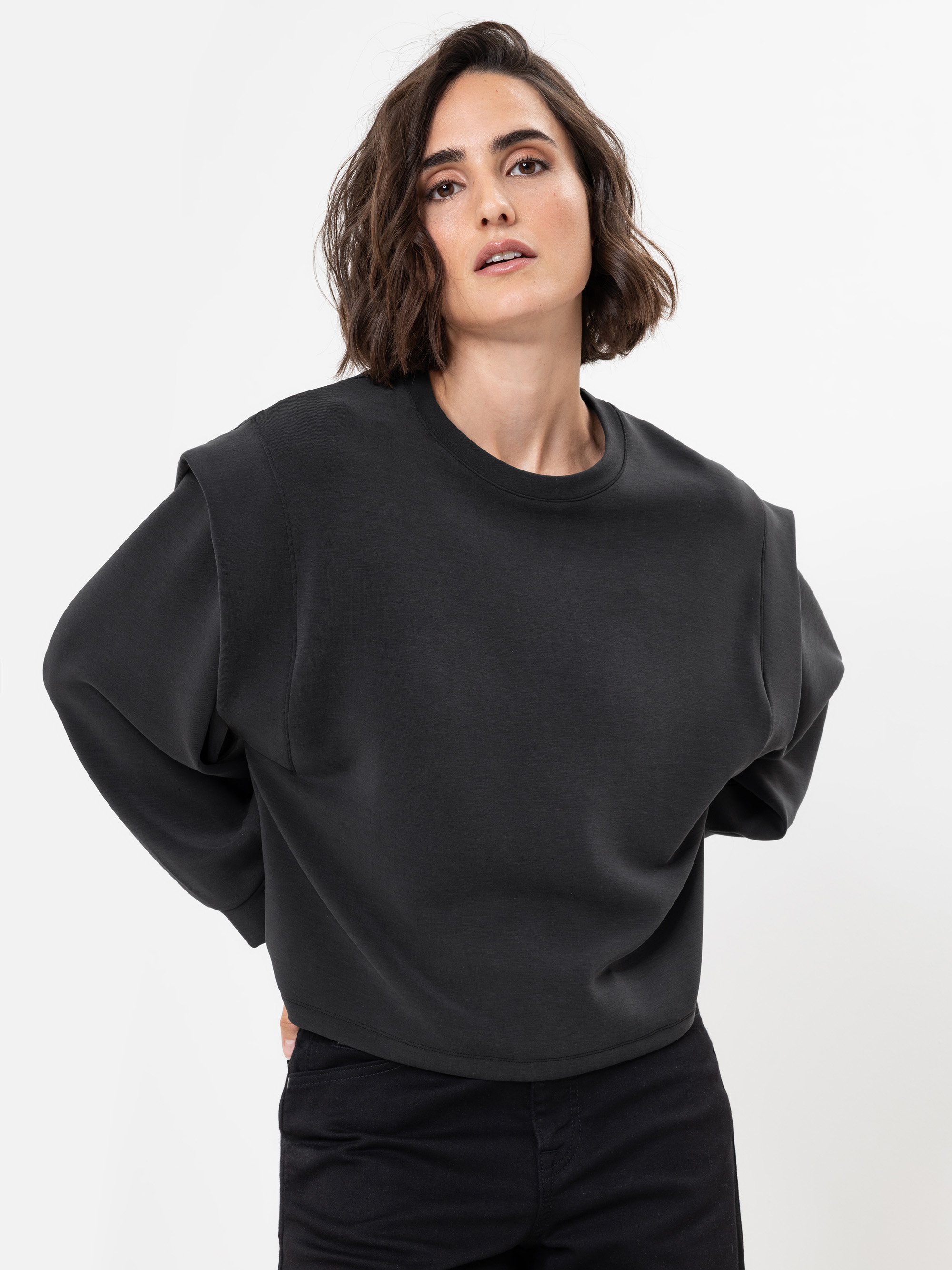 Eine Frau mit kurzen braunen Haaren, die ein locker sitzendes schwarzes Sweatshirt und eine schwarze Hose trägt, steht vor einem schlichten weißen Hintergrund und blickt selbstbewusst mit den Händen auf dem Rücken in die Kamera.