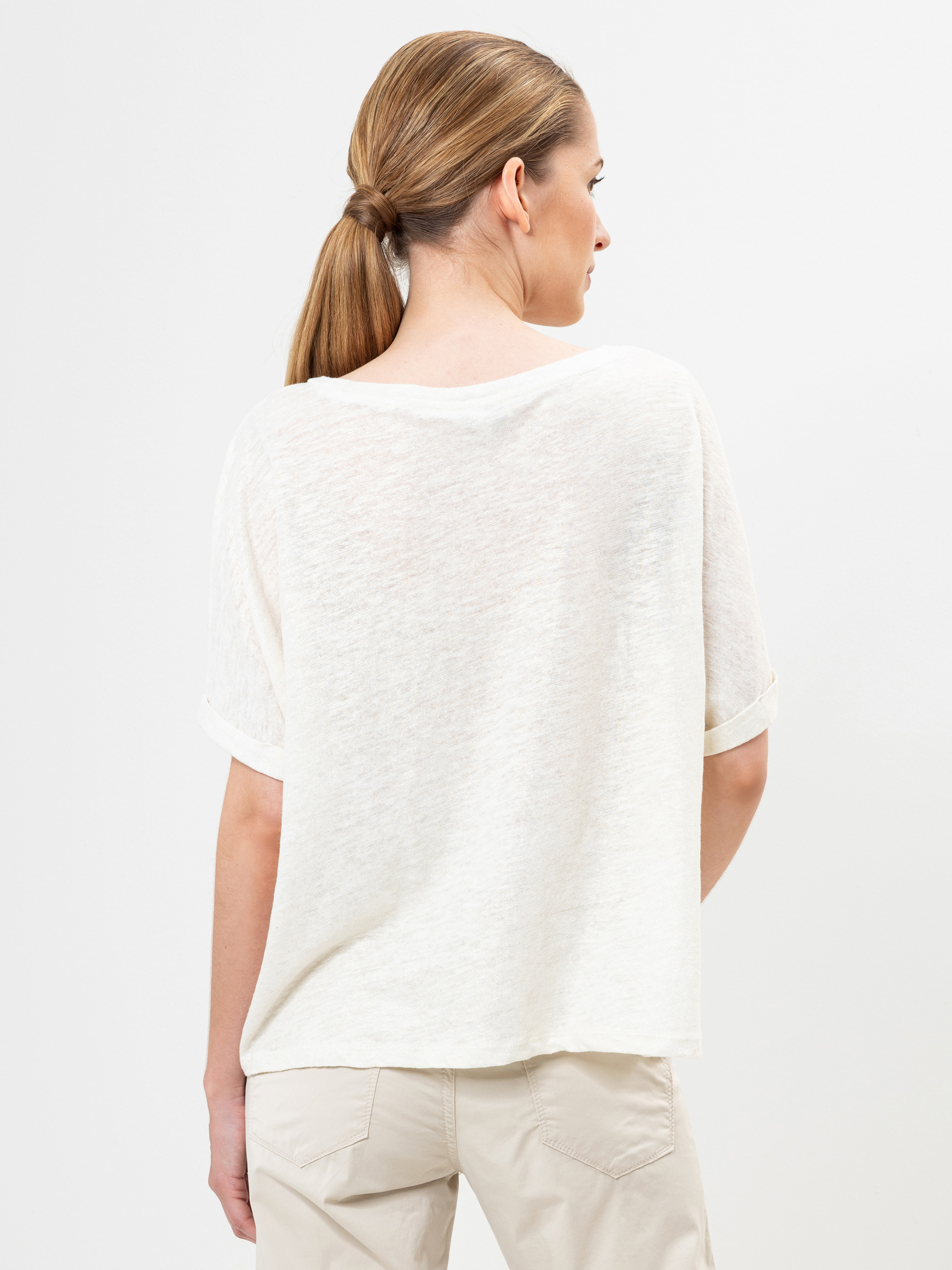 Eine Frau mit hellbraunem Haar in einem niedrigen Pferdeschwanz wird von hinten gezeigt. Sie trägt ein locker sitzendes, kurzärmeliges, cremefarbenes Oberteil und eine beigefarbene Hose vor einem schlichten weißen Hintergrund.