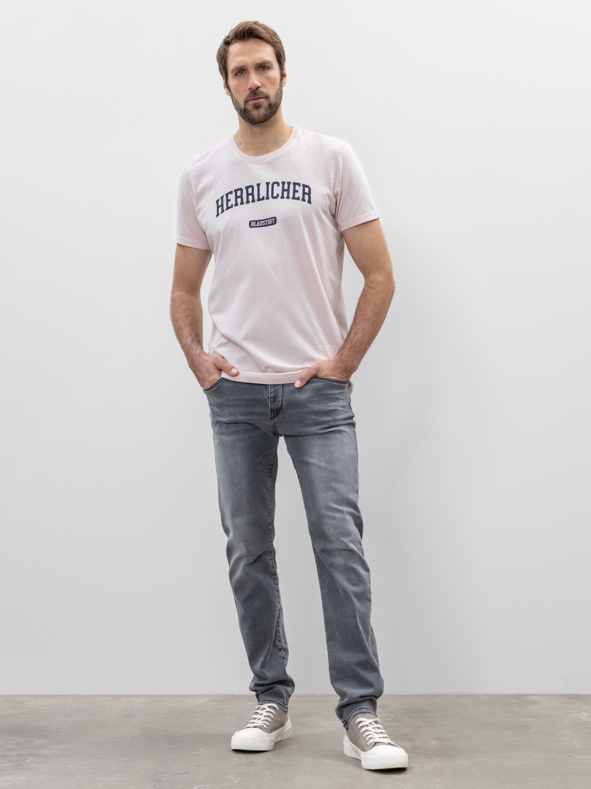 Ein Mann steht vor einer schlichten hellgrauen Wand. Er trägt ein hellrosa T-Shirt mit dem Aufdruck HERALICHER, graue Jeans und graue Turnschuhe. Er hat kurze Haare und einen Bart, die Hände stecken in den Taschen seiner Jeans.