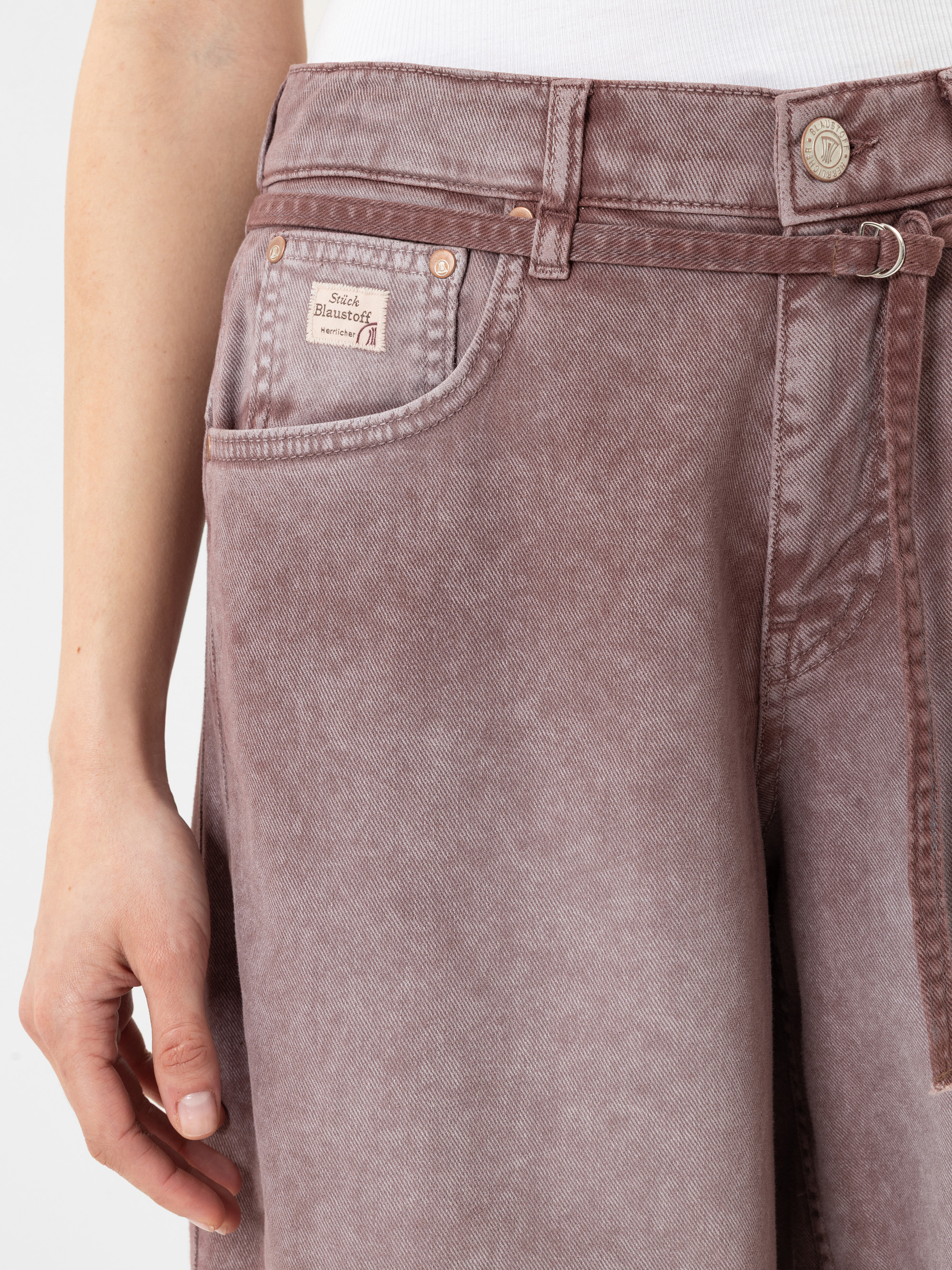 Eine Person trägt eine lilafarbene Jeans mit hoher Taille, einen passenden Gürtel und ein weißes Oberteil. Auf der Tasche der Jeans befindet sich ein kleines Etikett mit der Aufschrift Studio Massimo Rebecchi. Nur der untere Teil des Oberkörpers und d