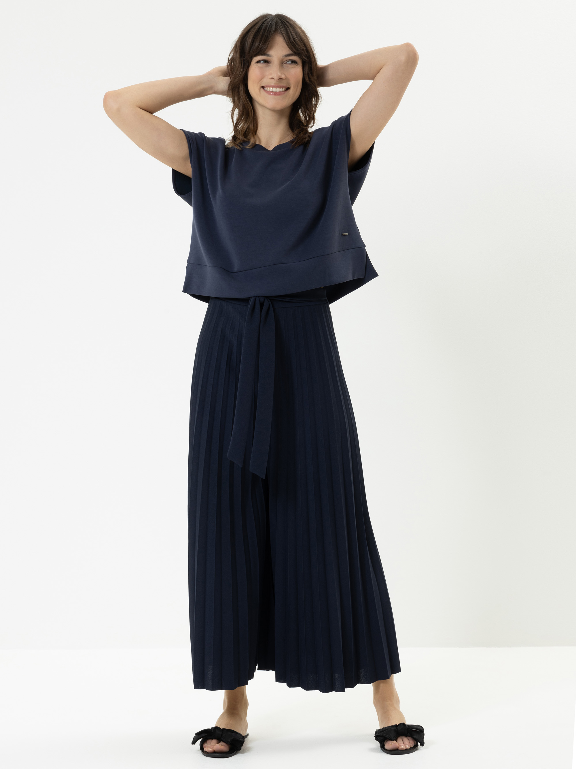 Eine lächelnde Frau steht mit den Händen hinter dem Kopf, trägt ein lockeres marineblaues Oberteil und eine dazu passende, in Falten gelegte Hose mit weitem Bein, die in der Taille gebunden ist, sowie schwarze Sandalen vor einem schlichten weißen Hint