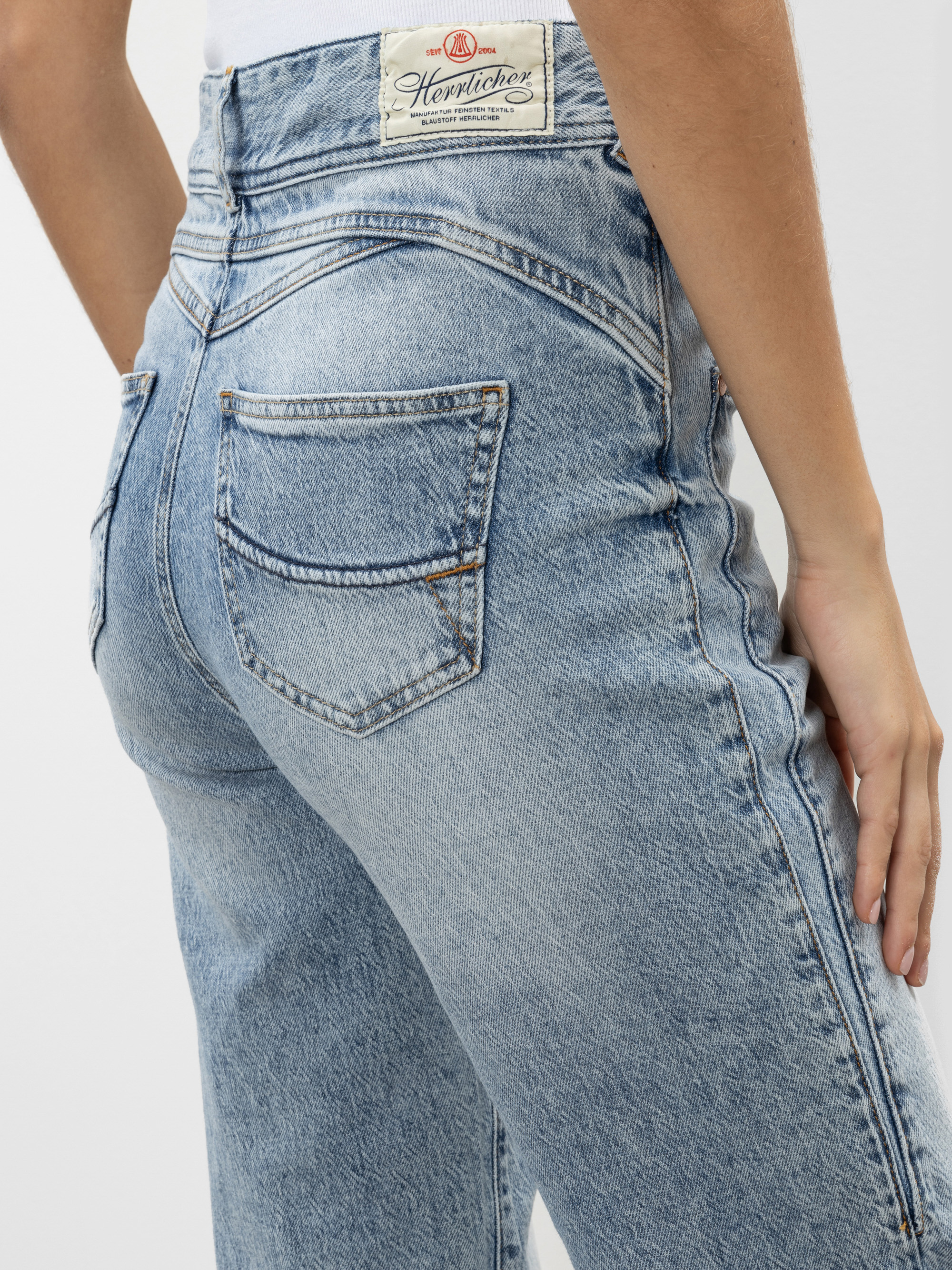 Eine Person trägt eine hellblaue Jeans mit hoher Taille, die von hinten und von der Taille abwärts gezeigt wird. Die Jeans hat eine Gesäßtasche und ein weißes Etikett mit Text am Bund. Die Hand der Person ruht an ihrer Seite.