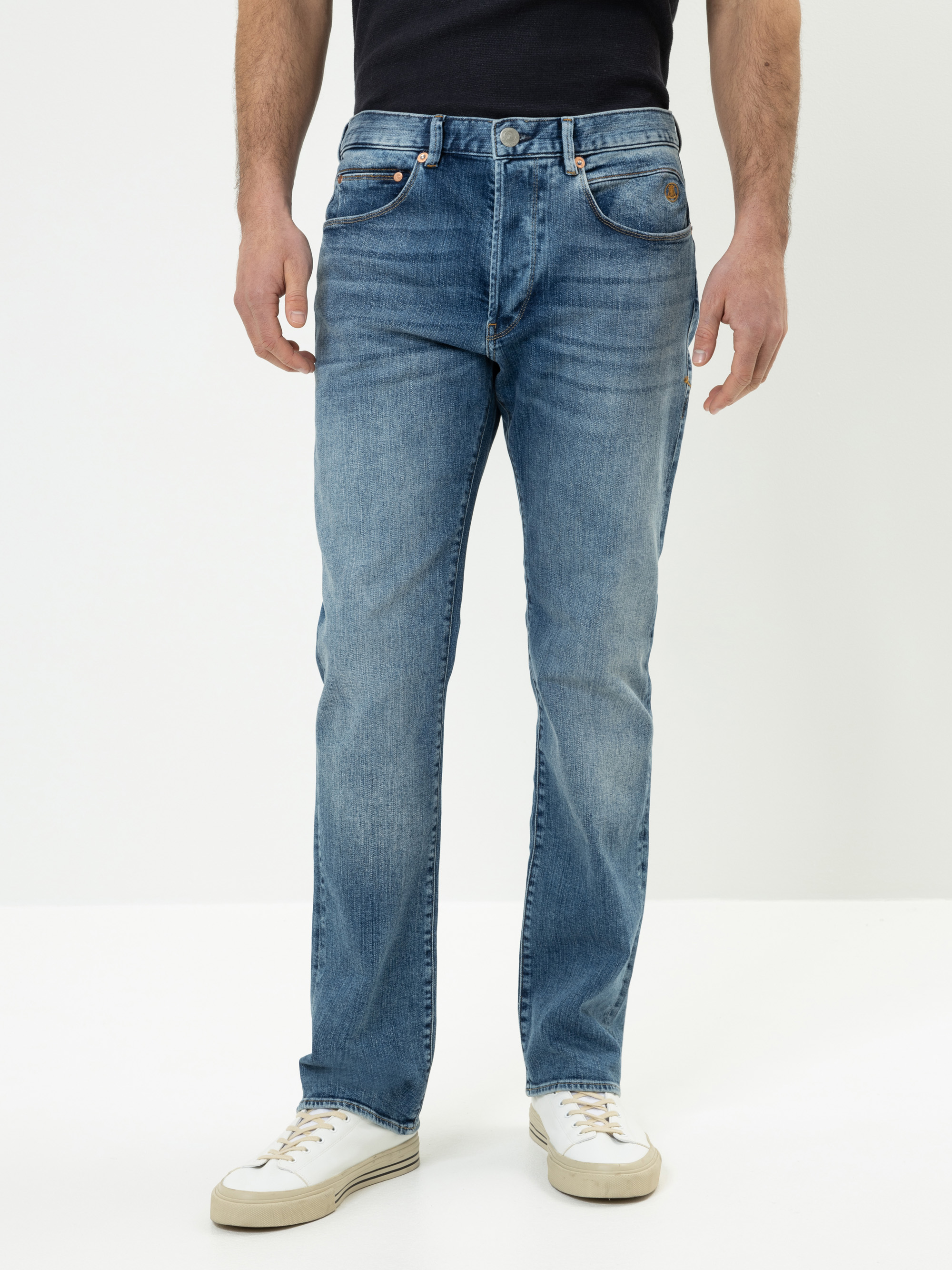 Eine Person, die eine hellblaue Jeans mit geradem Bein, ein dunkles Hemd und weiße Turnschuhe trägt, steht vor einem schlichten weißen Hintergrund, der von den Schultern bis zu den Füßen reicht.