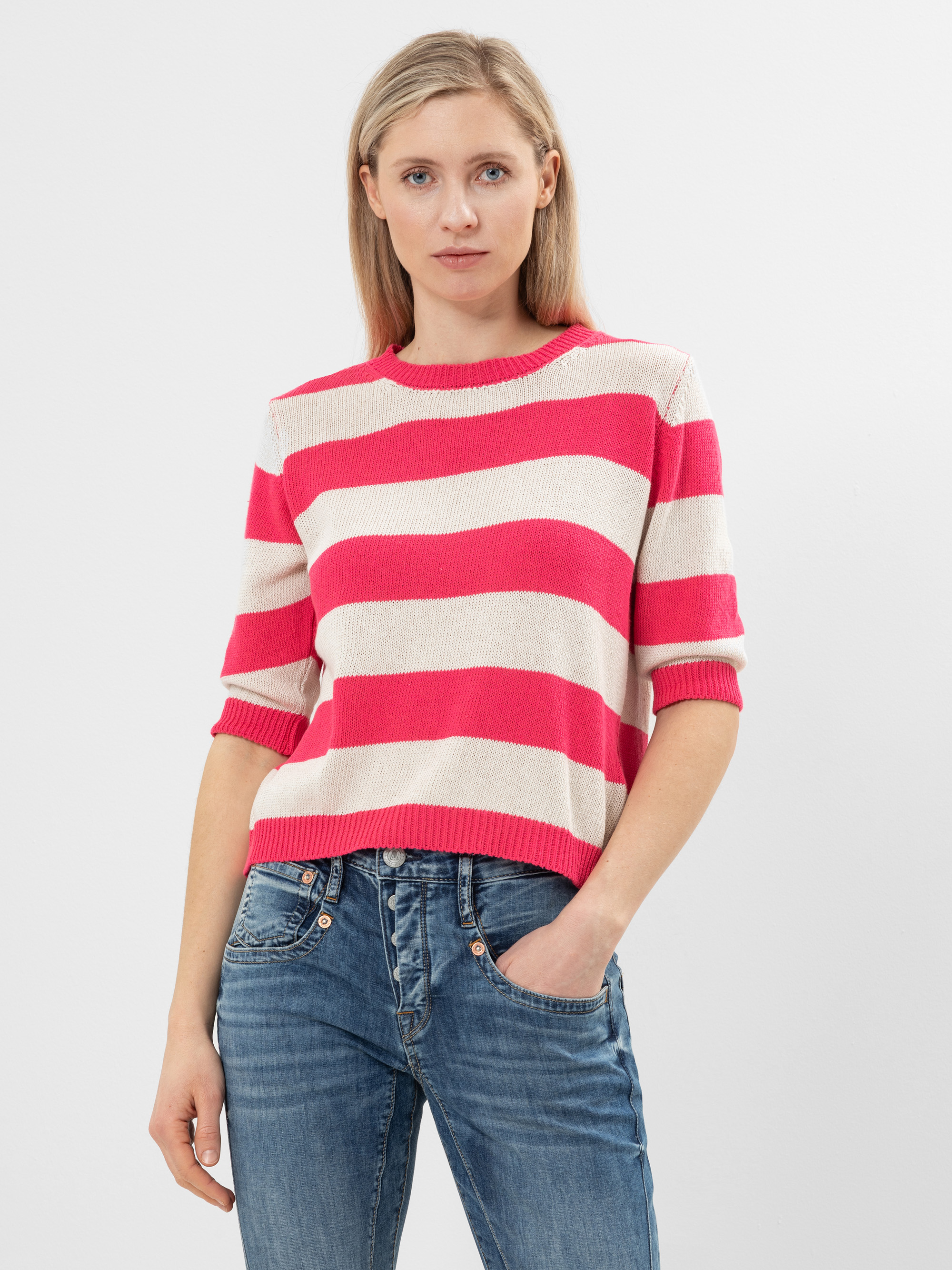 Eine Person mit langen blonden Haaren trägt einen rosa-weiß gestreiften Pullover und blaue Jeans und steht mit neutralem Gesichtsausdruck vor einem schlichten weißen Hintergrund.