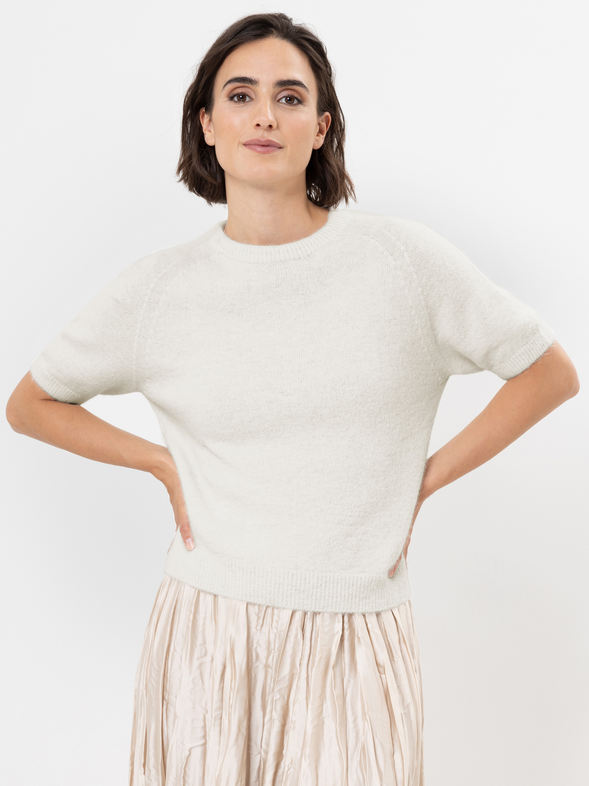 Eine Frau mit kurzen braunen Haaren, die einen cremefarbenen kurzärmeligen Pullover und einen leichten Faltenrock trägt, steht mit den Händen in den Hüften vor einem schlichten weißen Hintergrund und schaut selbstbewusst in die Kamera.