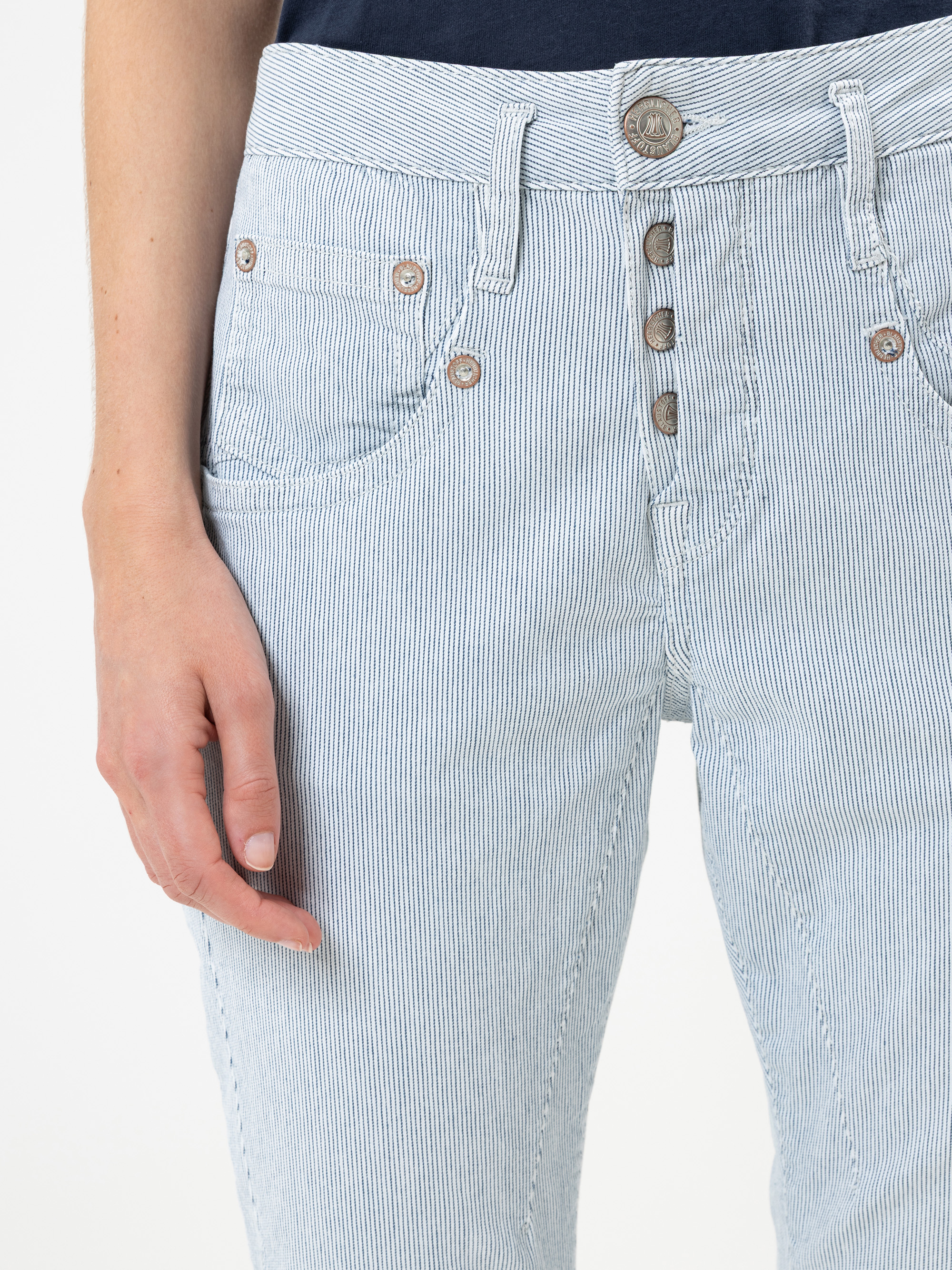 Eine Person trägt eine hellblau-weiß gestreifte Jeans mit mehreren sichtbaren Knöpfen. Nur der untere Teil des Oberkörpers und die linke Hand sind zu sehen. Die Hand liegt entspannt an der Seite. Der Hintergrund ist einfarbig weiß.
