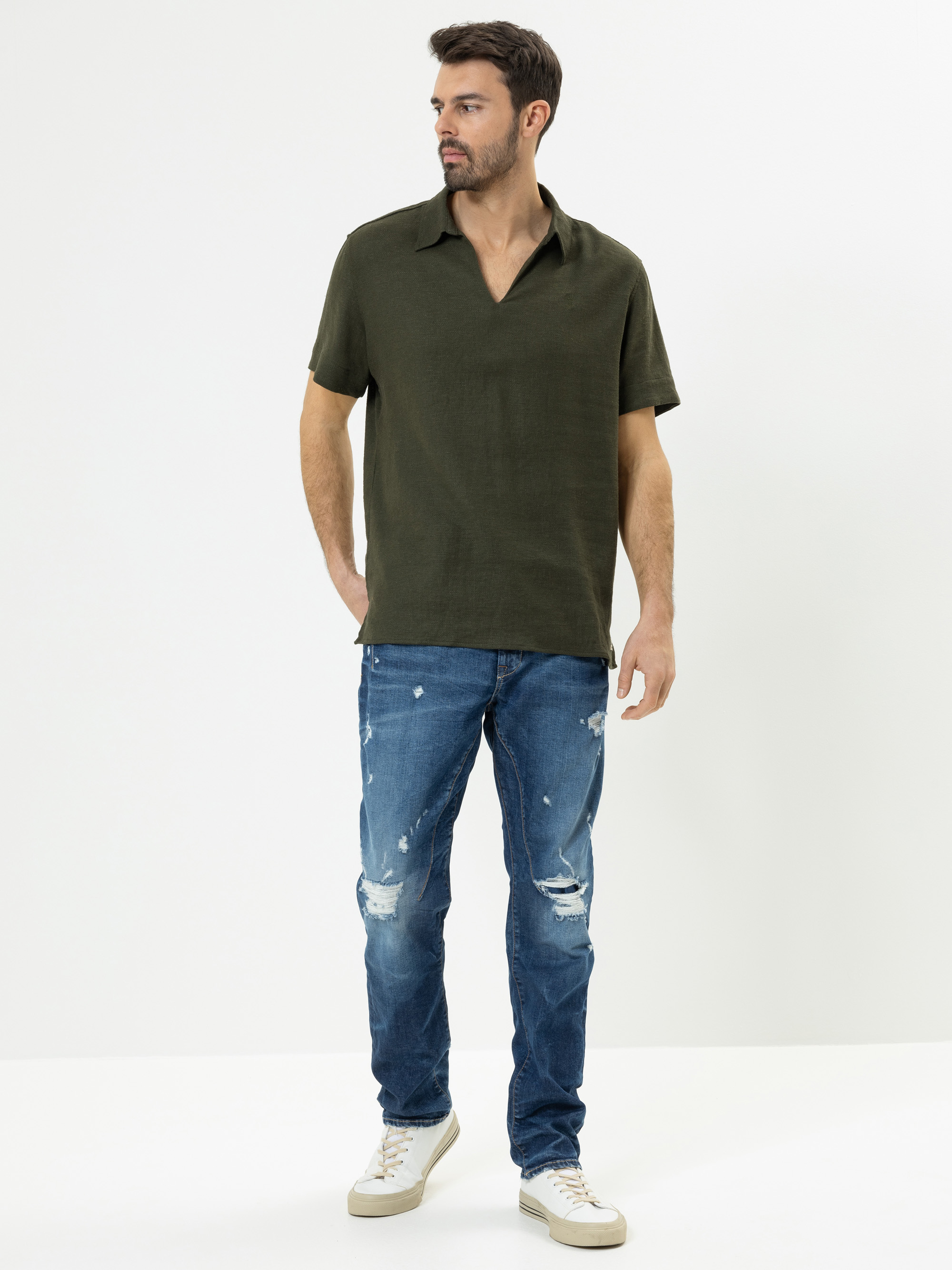 Ein Mann steht vor einem schlichten weißen Hintergrund und trägt ein dunkelgrünes, kurzärmeliges Hemd, abgewetzte Bluejeans und weiße Turnschuhe. Er blickt nach links und hat eine Hand in der Tasche.