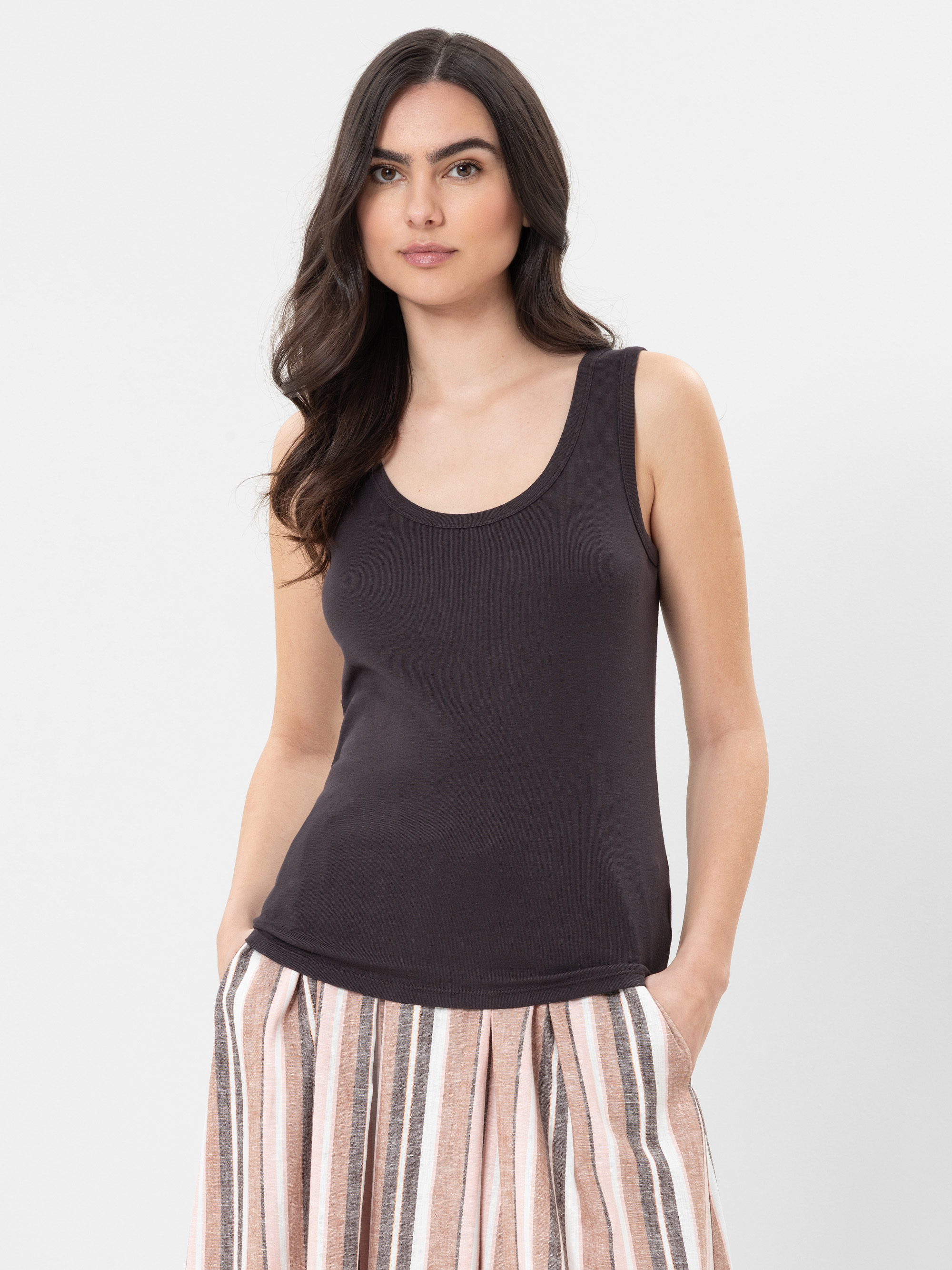 Eine Frau mit langen dunklen Haaren, die ein ärmelloses schwarzes Oberteil und einen beige, rosa und braun gestreiften Rock trägt, steht mit den Händen in den Taschen vor einem schlichten hellen Hintergrund.