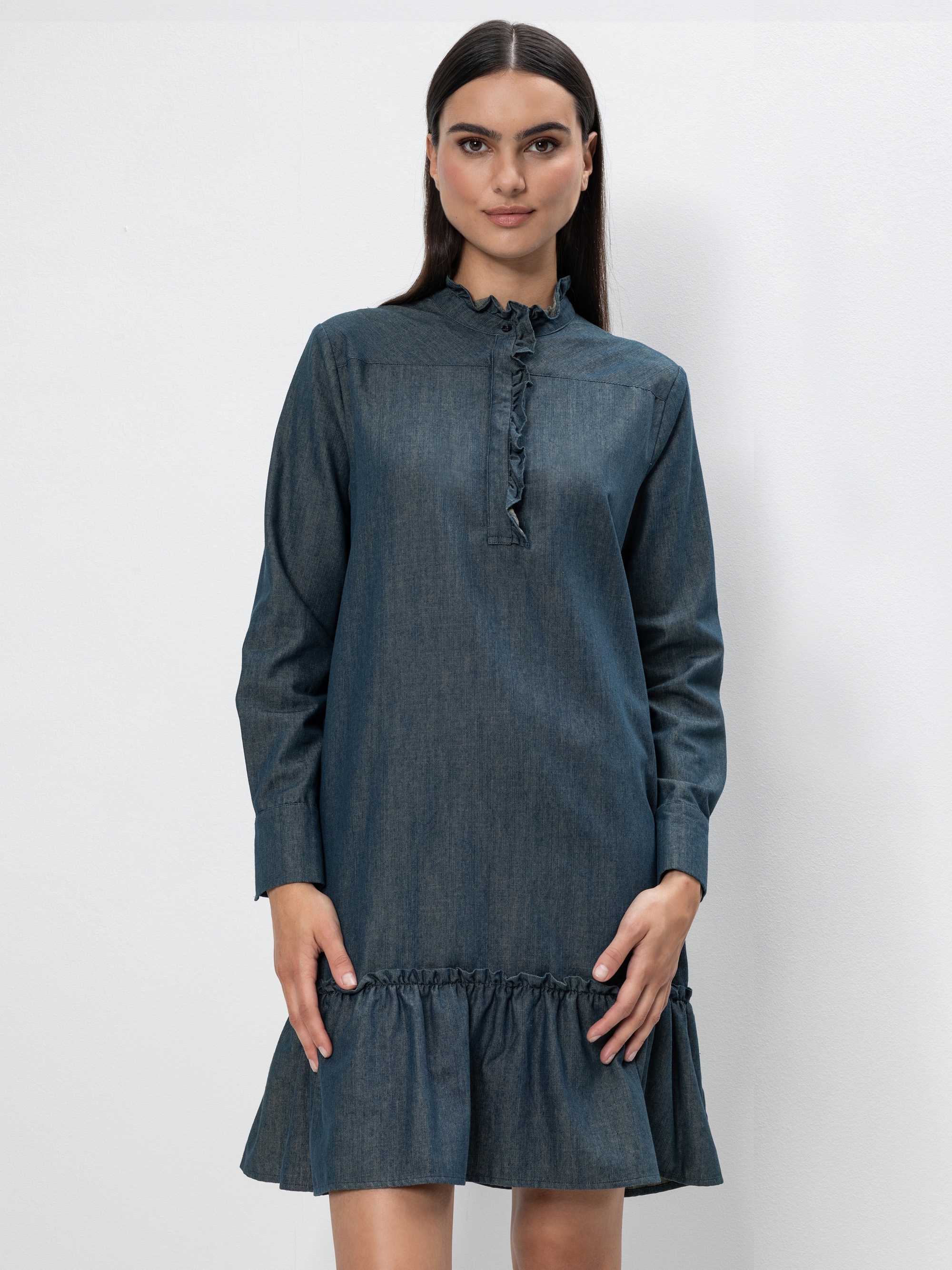 Eine Frau mit langen dunklen Haaren trägt ein blaues, langärmeliges Jeanskleid mit einem gerüschten Kragen, einer Knopfleiste auf der Vorderseite und einem gerüschten Saum vor einem schlichten weißen Hintergrund.