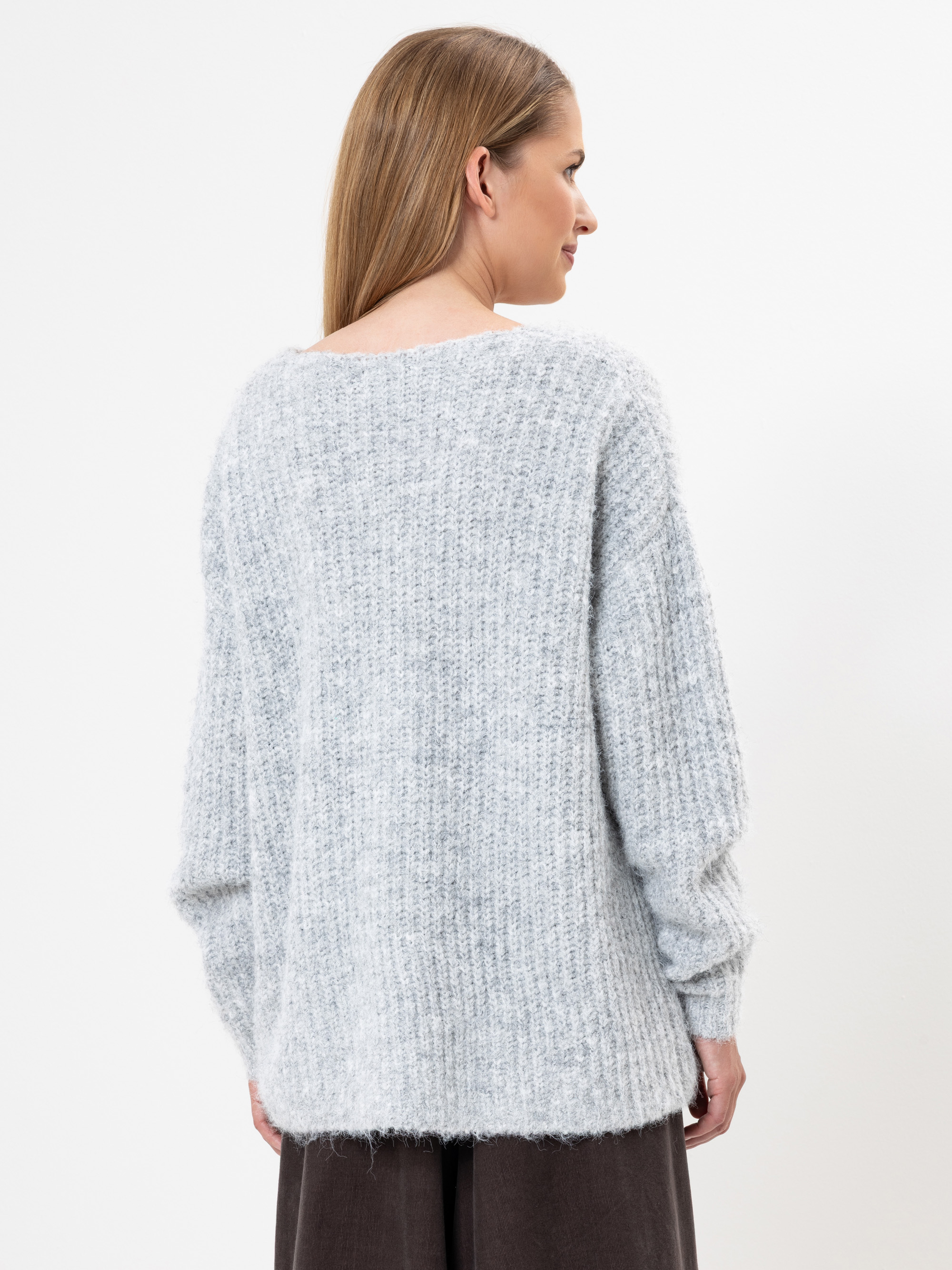 Eine Frau mit glattem, hellbraunem Haar trägt einen lockeren, hellgrauen, strukturierten Strickpullover und eine dunkle Hose. Sie steht vor einem schlichten weißen Hintergrund und blickt von der Kamera weg.