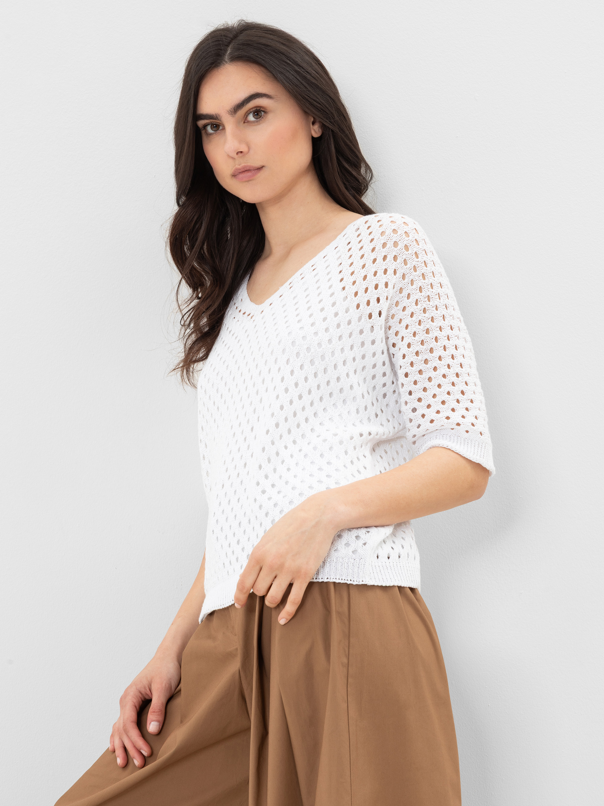 Eine Frau mit langen braunen Haaren trägt einen weißen, kurzärmeligen Strickpullover mit V-Ausschnitt und eine hellbraune, weit geschnittene Hose. Sie steht vor einem schlichten, hellen Hintergrund und blickt leicht zur Seite.