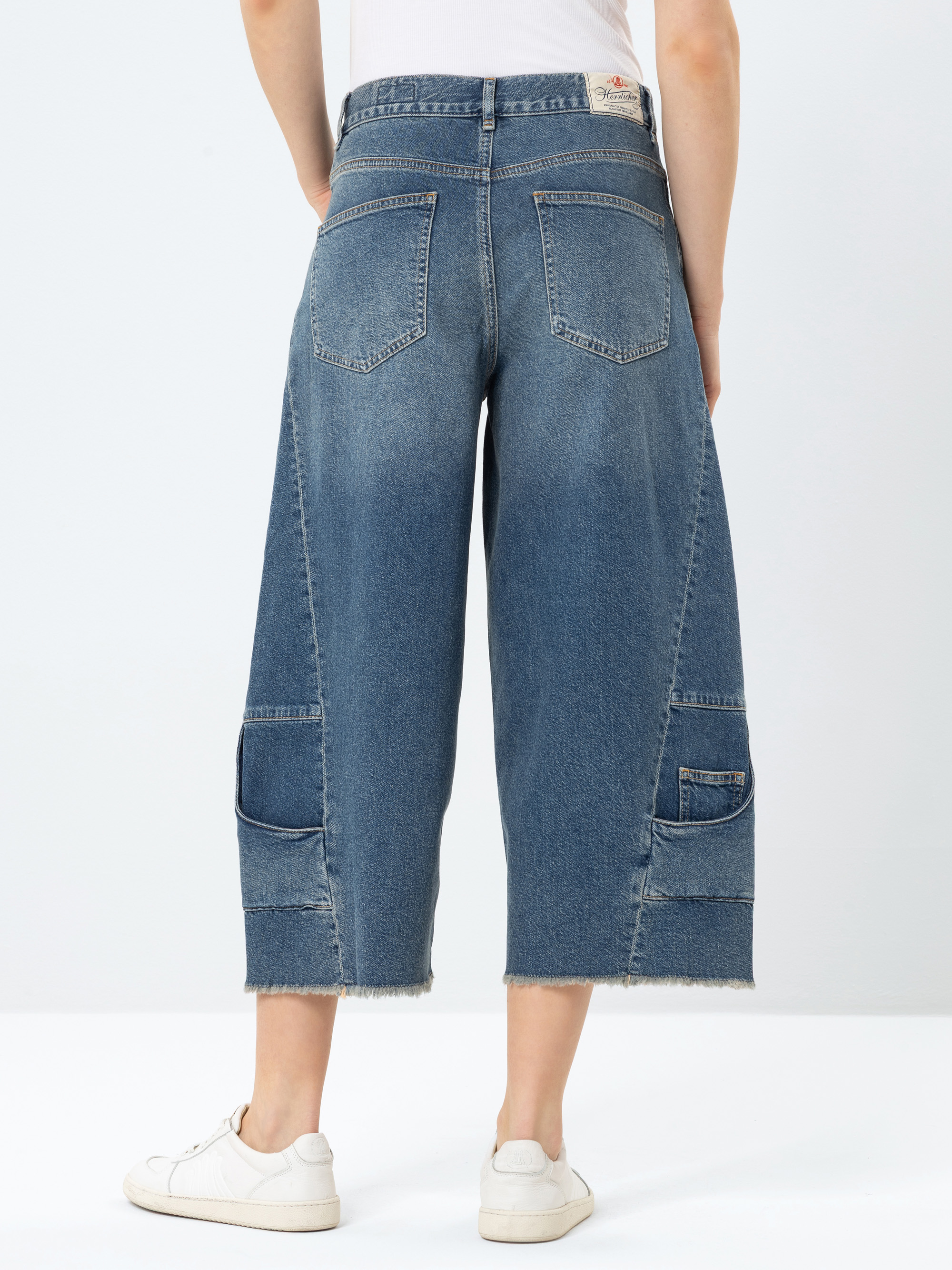 Person, die eine blaue Jeans mit weitem Bein, rohem Saum, großen Gesäßtaschen und zusätzlichen aufgesetzten Taschen in der Nähe der Knie trägt, gepaart mit weißen Turnschuhen, von hinten vor einem unifarbenen Hintergrund gezeigt.