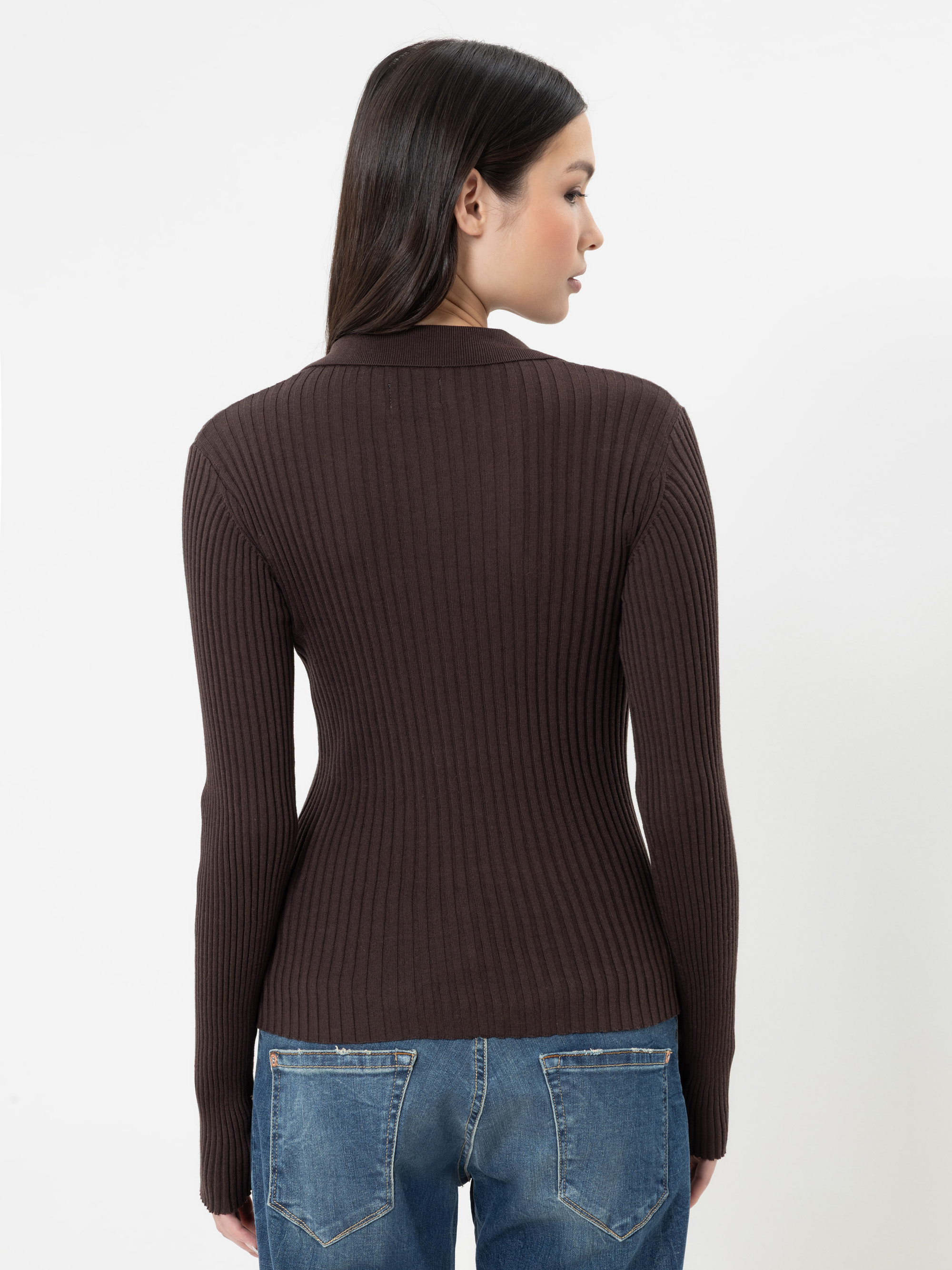 Eine Frau mit langen dunklen Haaren, die einen eng anliegenden, gerippten, dunkelbraunen Pullover und eine blaue Jeans trägt, steht mit dem Rücken zur Kamera vor einem schlichten weißen Hintergrund.