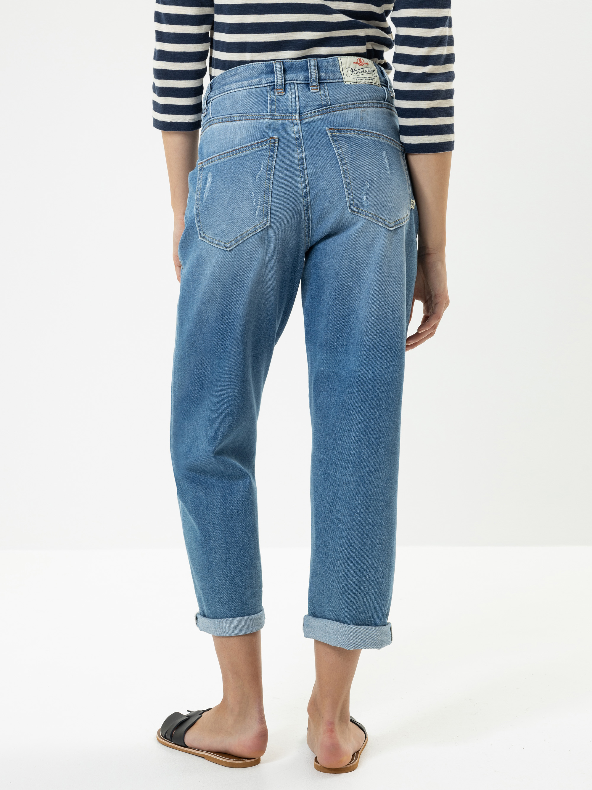 Eine Person steht barfuß mit dem Gesicht nach außen und trägt eine hellblaue Jeans mit Bündchen und ein marineblau-weiß gestreiftes langärmeliges Hemd. Die Jeans hat Gesäßtaschen und eine lockere Passform. Der Hintergrund ist einfarbig weiß.