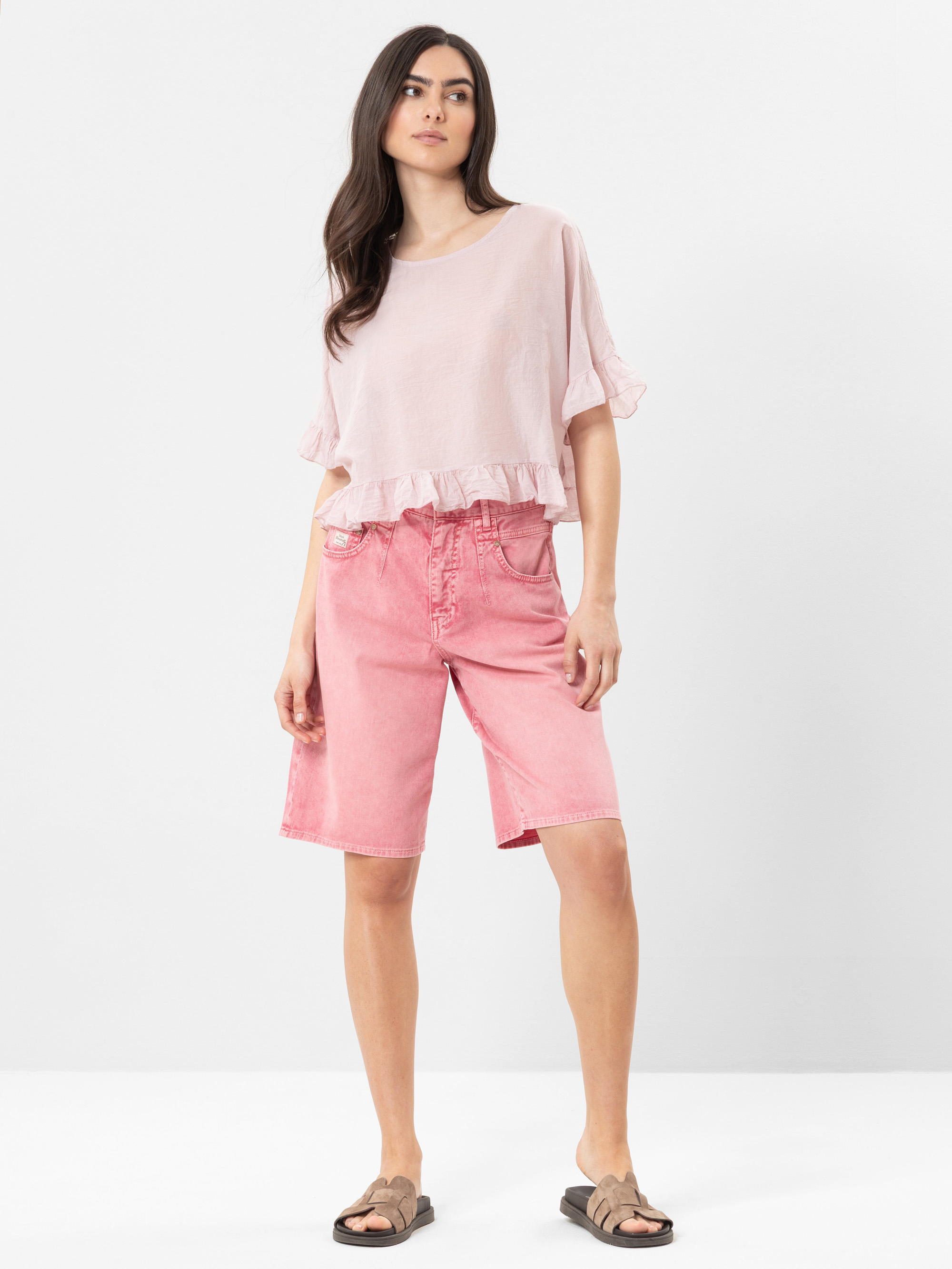 Eine Frau steht vor einem schlichten Hintergrund und trägt ein lockeres, hellrosa gerafftes Oberteil, knielange rosa Shorts und braune Sandalen. Sie hat langes dunkles Haar und einen neutralen Gesichtsausdruck.
