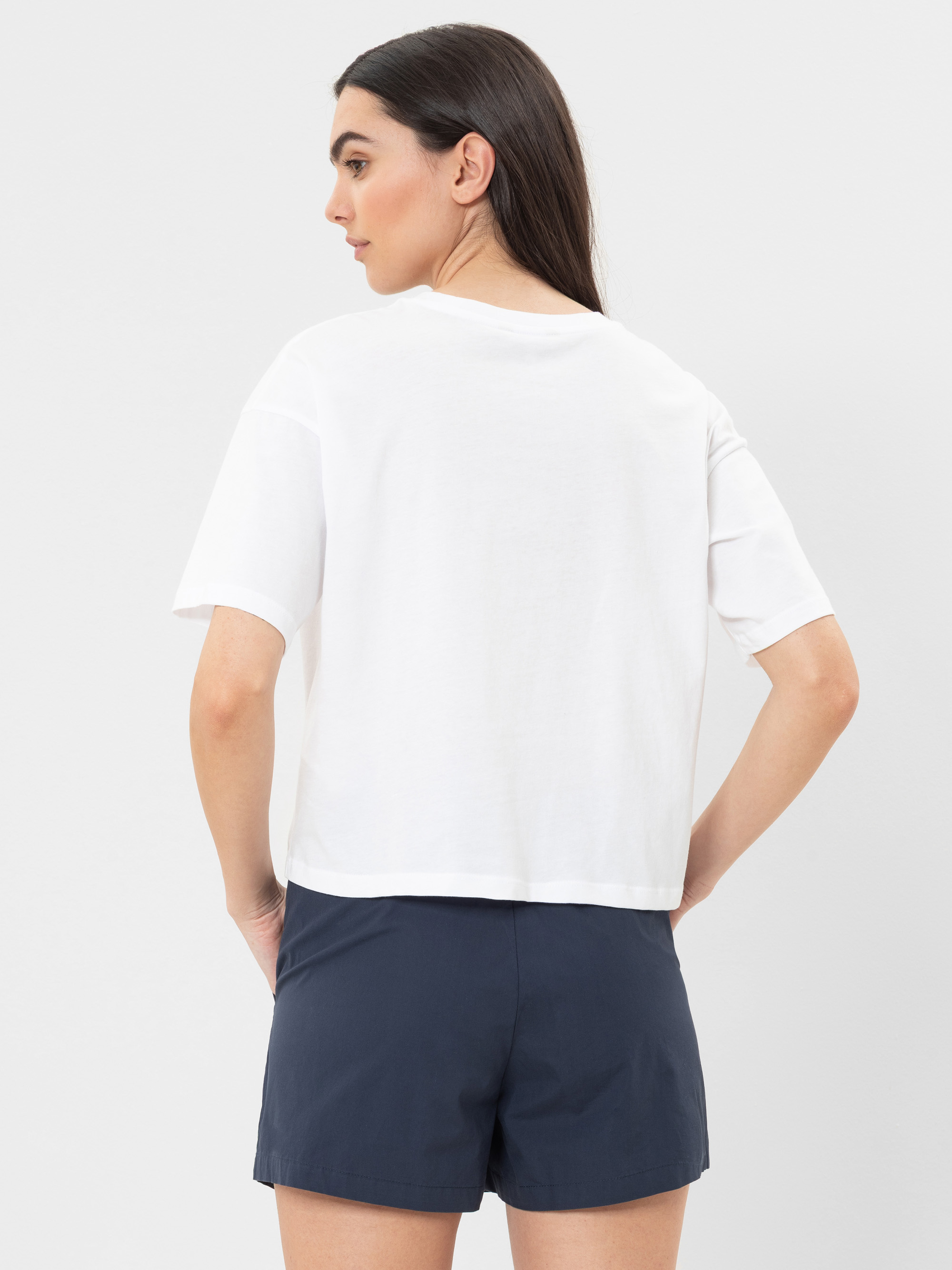 Eine Frau mit langen dunklen Haaren steht mit dem Gesicht zur Wand, trägt ein lockeres weißes T-Shirt und dunkle marineblaue Shorts vor einem schlichten hellen Hintergrund.