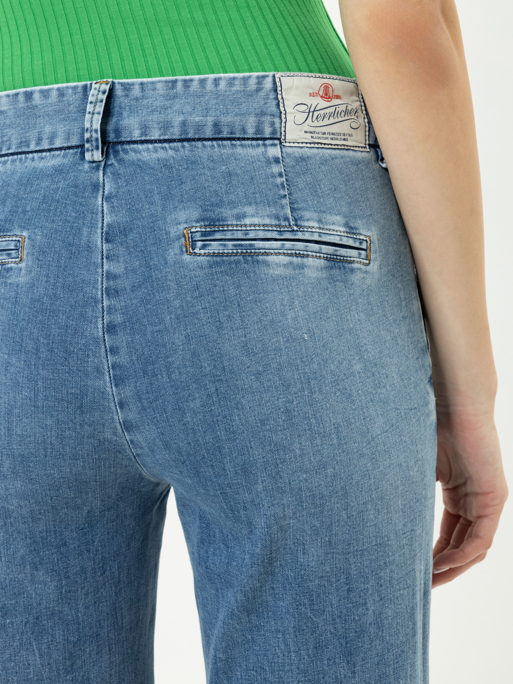 Gezeigt wird eine Person, die eine blaue Jeans und ein grünes geripptes Oberteil trägt, von der Taille abwärts, mit einer Nahaufnahme des hinteren Taschenbereichs und einem weißen Herviches-Etikett am Bund.