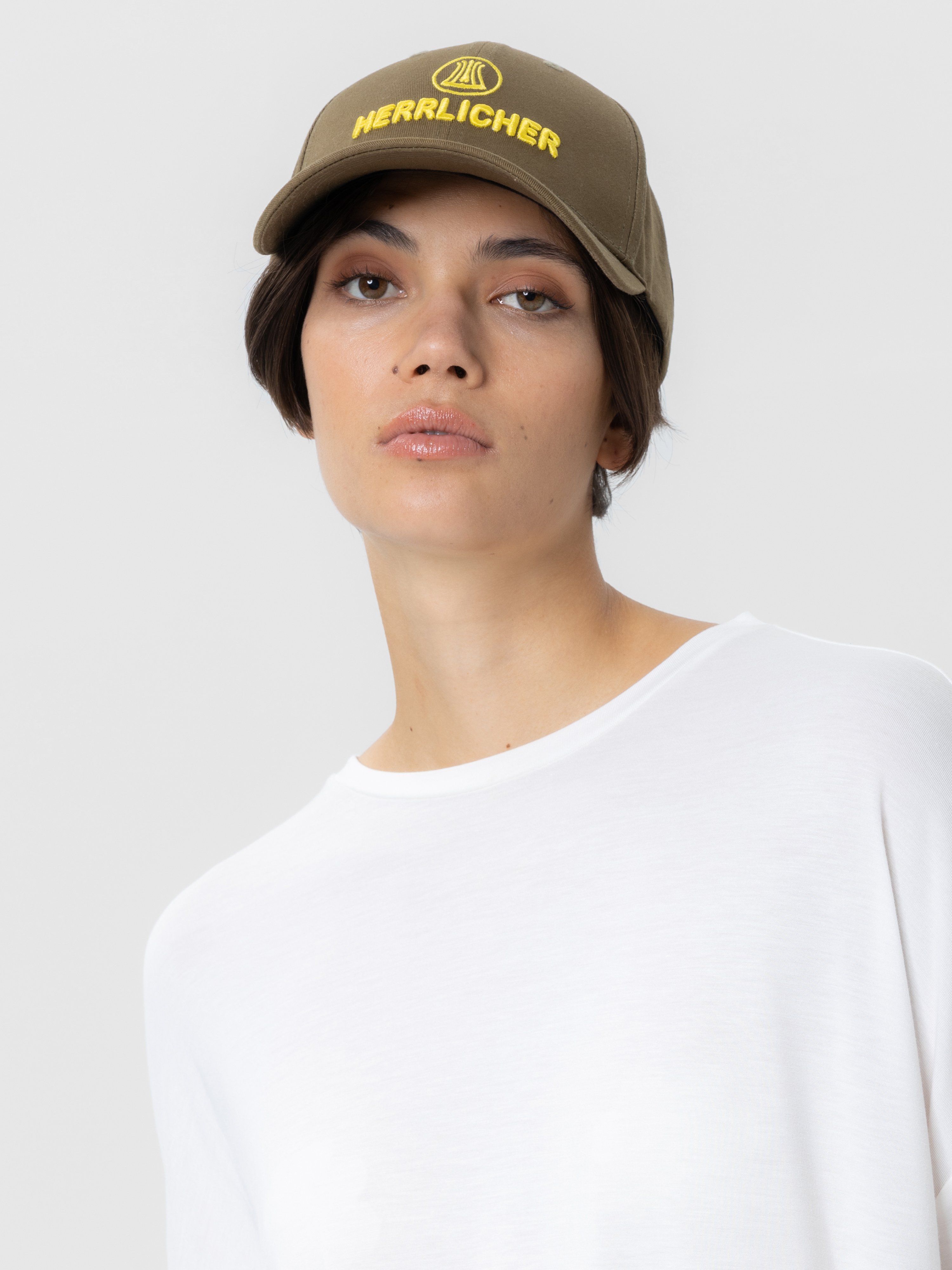 Eine Person, die eine olivgrüne Baseballkappe mit gelber Aufschrift HERRELICHER und ein weißes T-Shirt trägt, posiert vor einem schlichten hellgrauen Hintergrund.