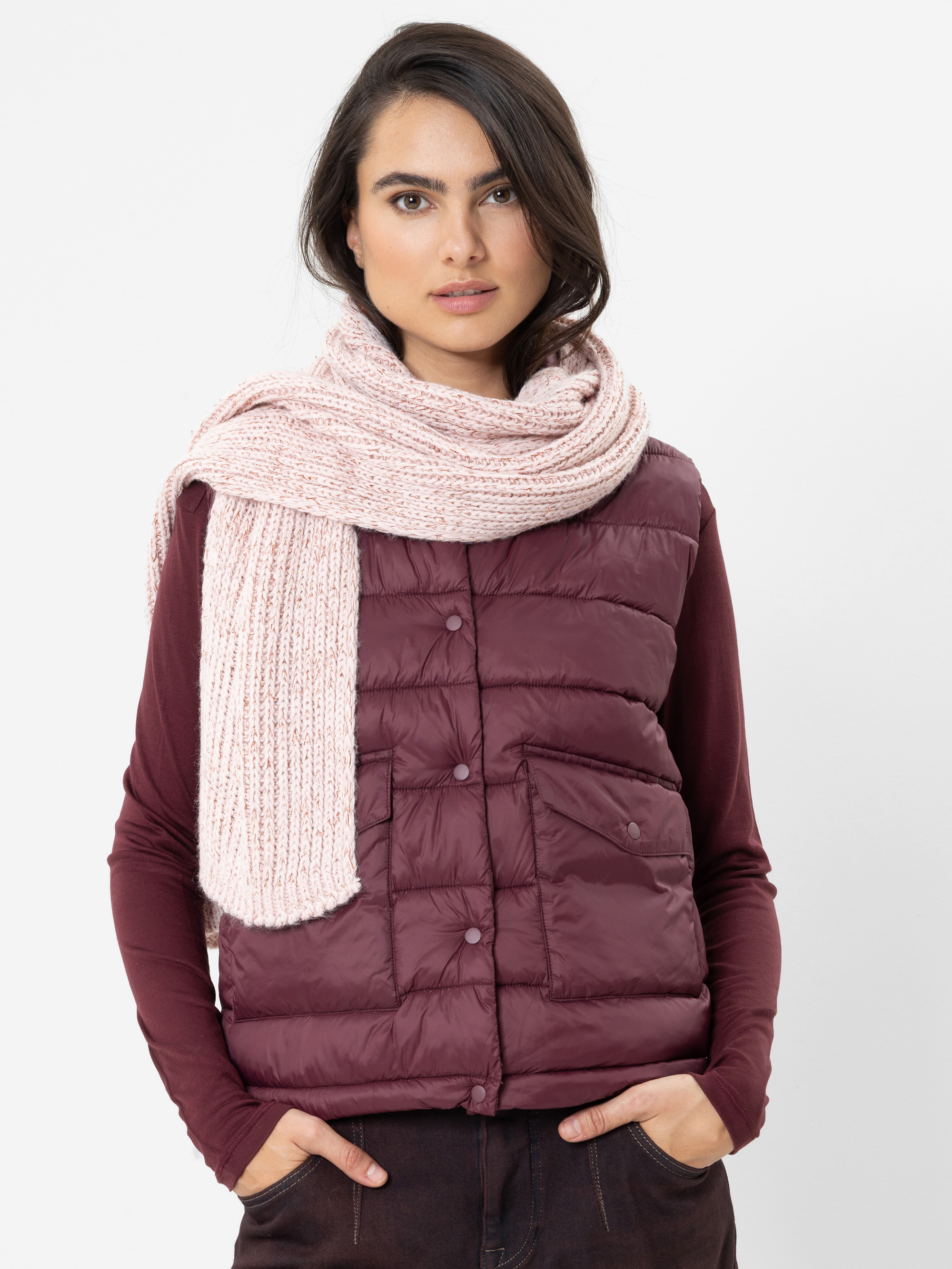Eine Frau mit langen dunklen Haaren, die eine weinrote Daunenweste, ein dazu passendes Langarmhemd und einen dicken, hellrosa Strickschal trägt und mit den Händen in den Taschen vor einem schlichten weißen Hintergrund steht.