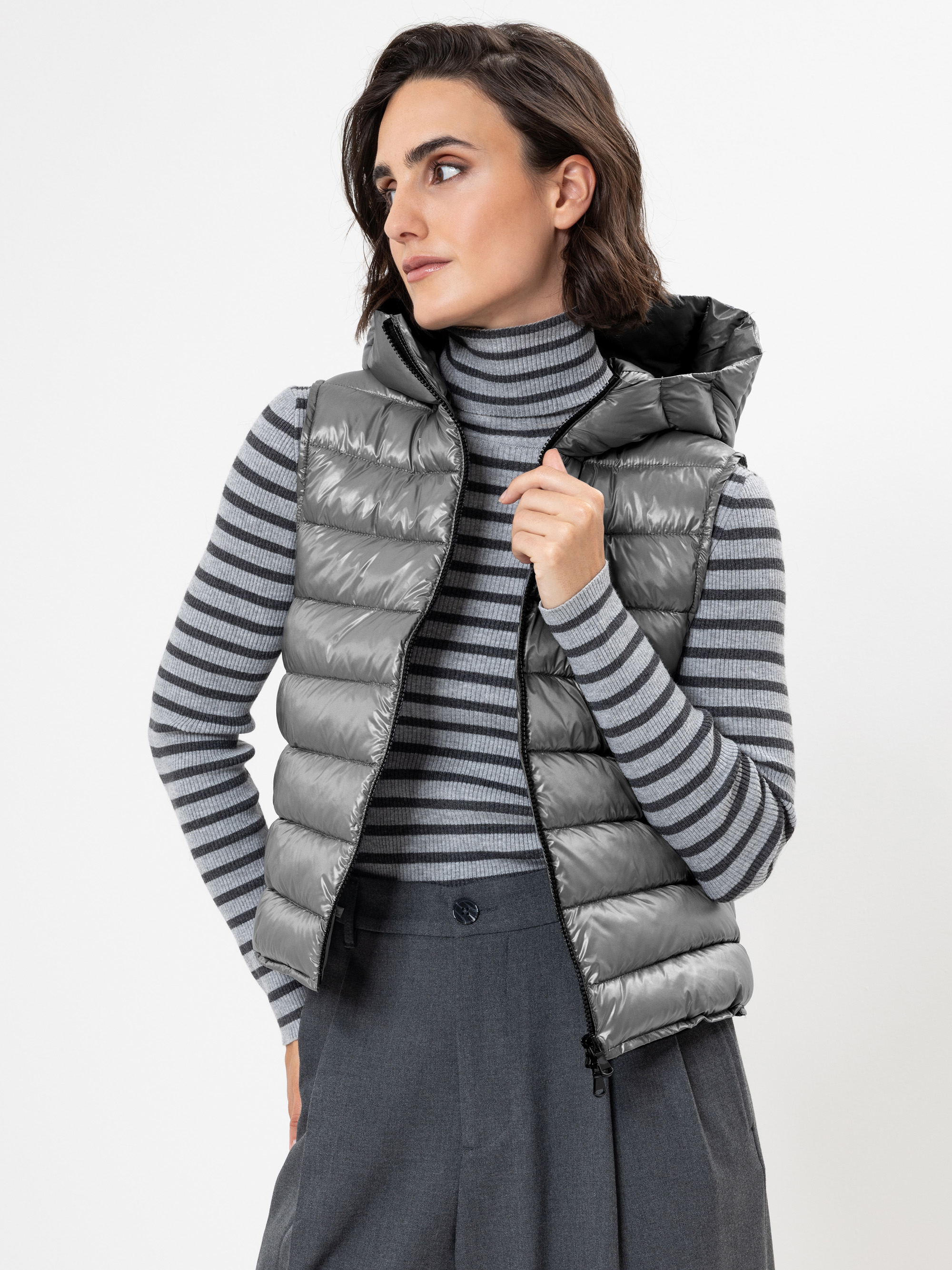 Eine Frau, die eine glänzende graue Pufferweste über einem grau-schwarz gestreiften Rollkragenpullover und eine graue Hose mit hoher Taille trägt, blickt vor einem schlichten weißen Hintergrund zur Seite.