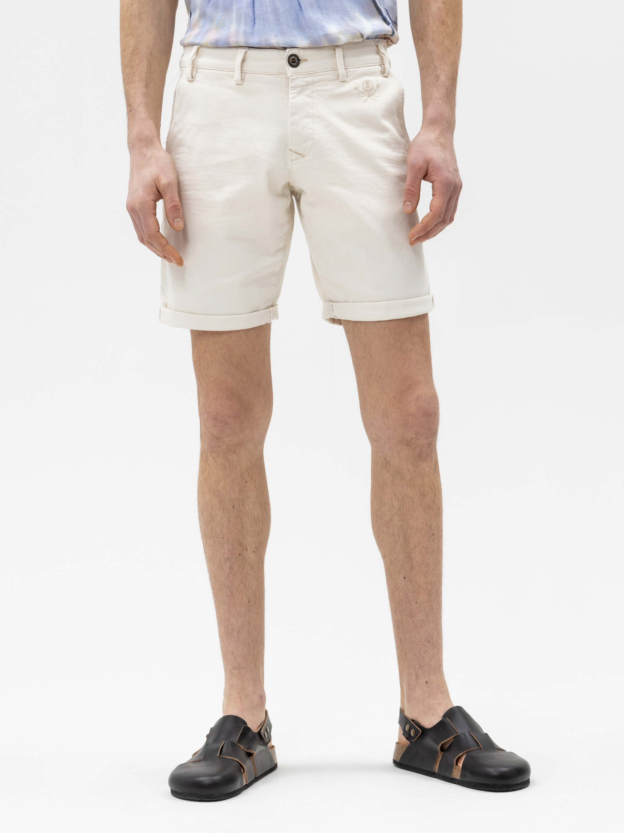 Eine Person, die vor einem weißen Hintergrund steht und cremefarbene, gekrempelte Shorts und schwarze Sandalen trägt. Nur die untere Hälfte des Körpers ist sichtbar.