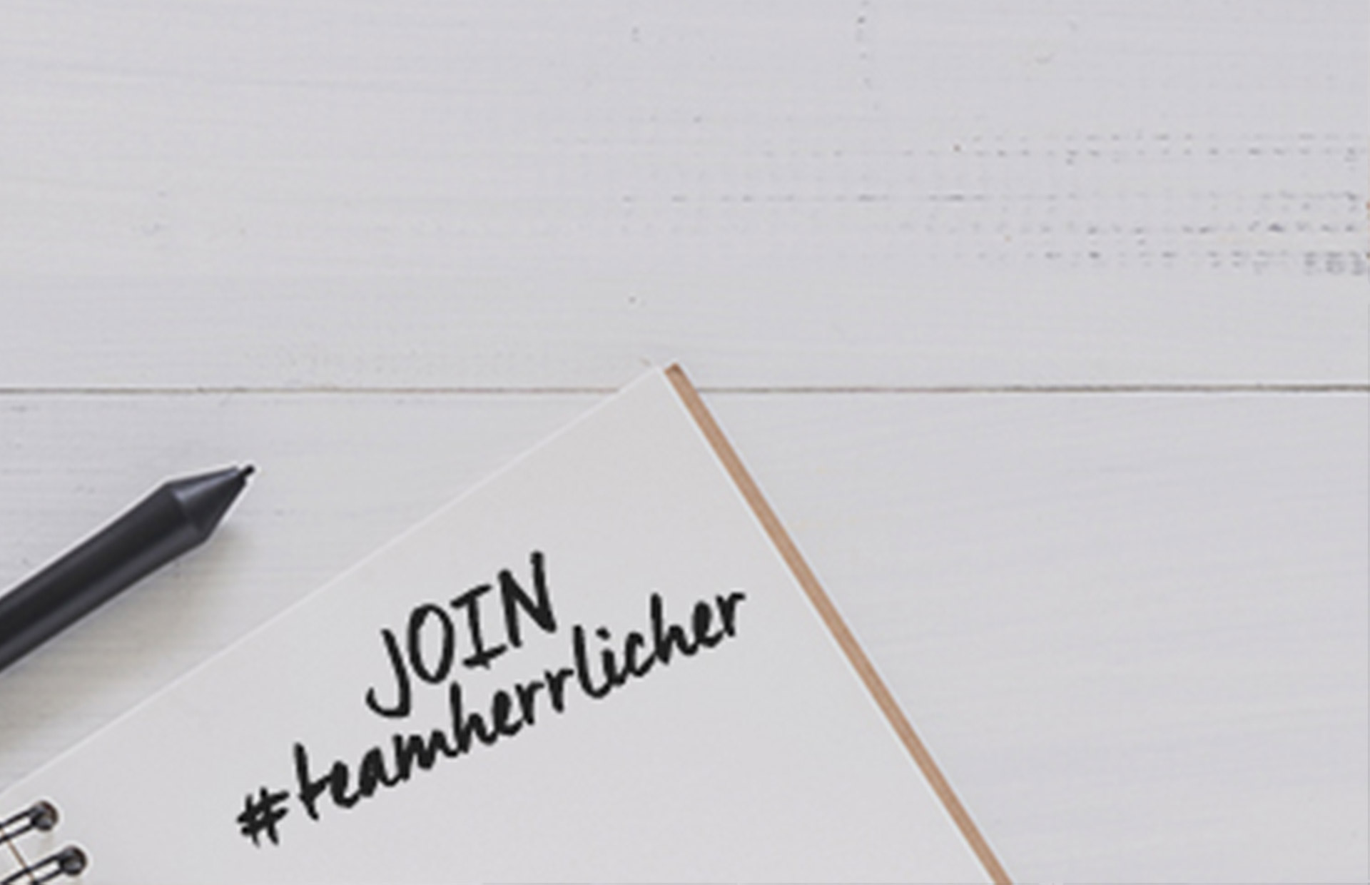 Ein schwarzer Stift liegt neben einem aufgeschlagenen Notizbuch mit den handgeschriebenen Worten JOIN #teamherrlicher auf einer weißen Holzoberfläche.