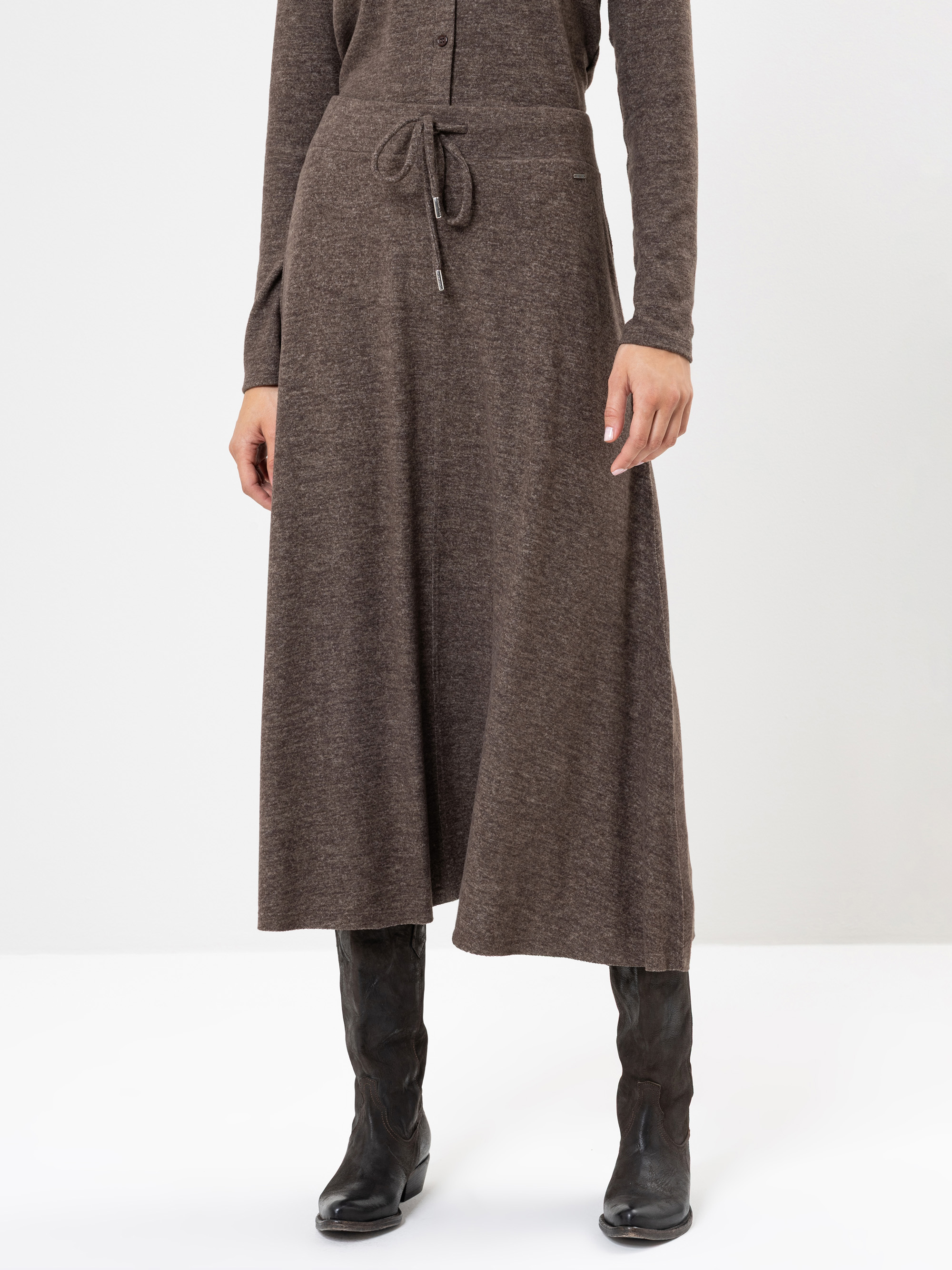 Eine Person, die einen langen, braunen Strickrock mit Kordelzug in der Taille und schwarze kniehohe Stiefel trägt, steht vor einem schlichten weißen Hintergrund. Der obere Teil des Rocks und die Unterarme sind sichtbar.