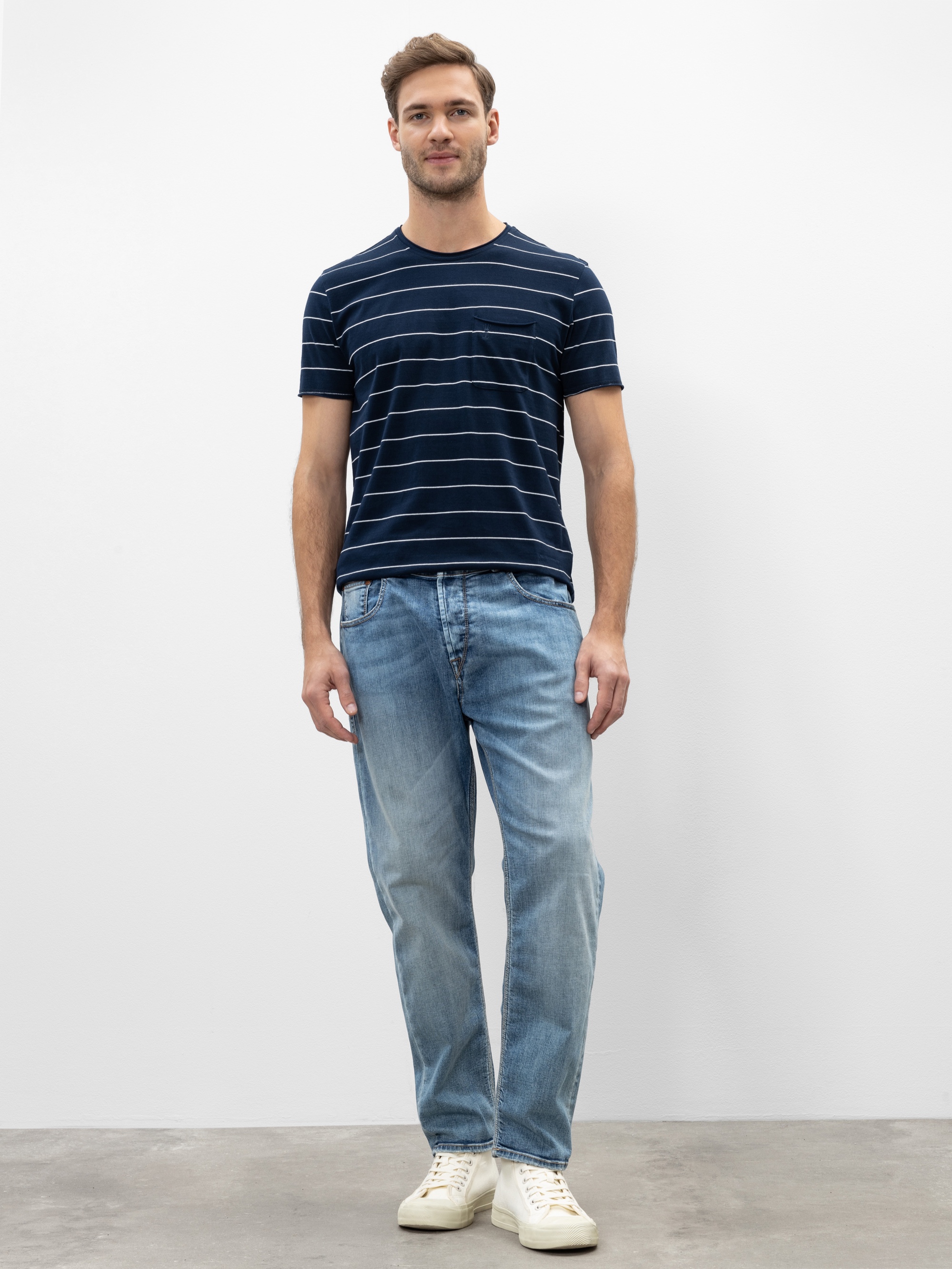 Ein Mann steht vor einer schlichten weißen Wand und trägt ein marineblau-weiß gestreiftes T-Shirt, hellblaue Jeans und weiße Turnschuhe. Er hat kurze Haare und schaut mit neutralem Blick direkt in die Kamera.