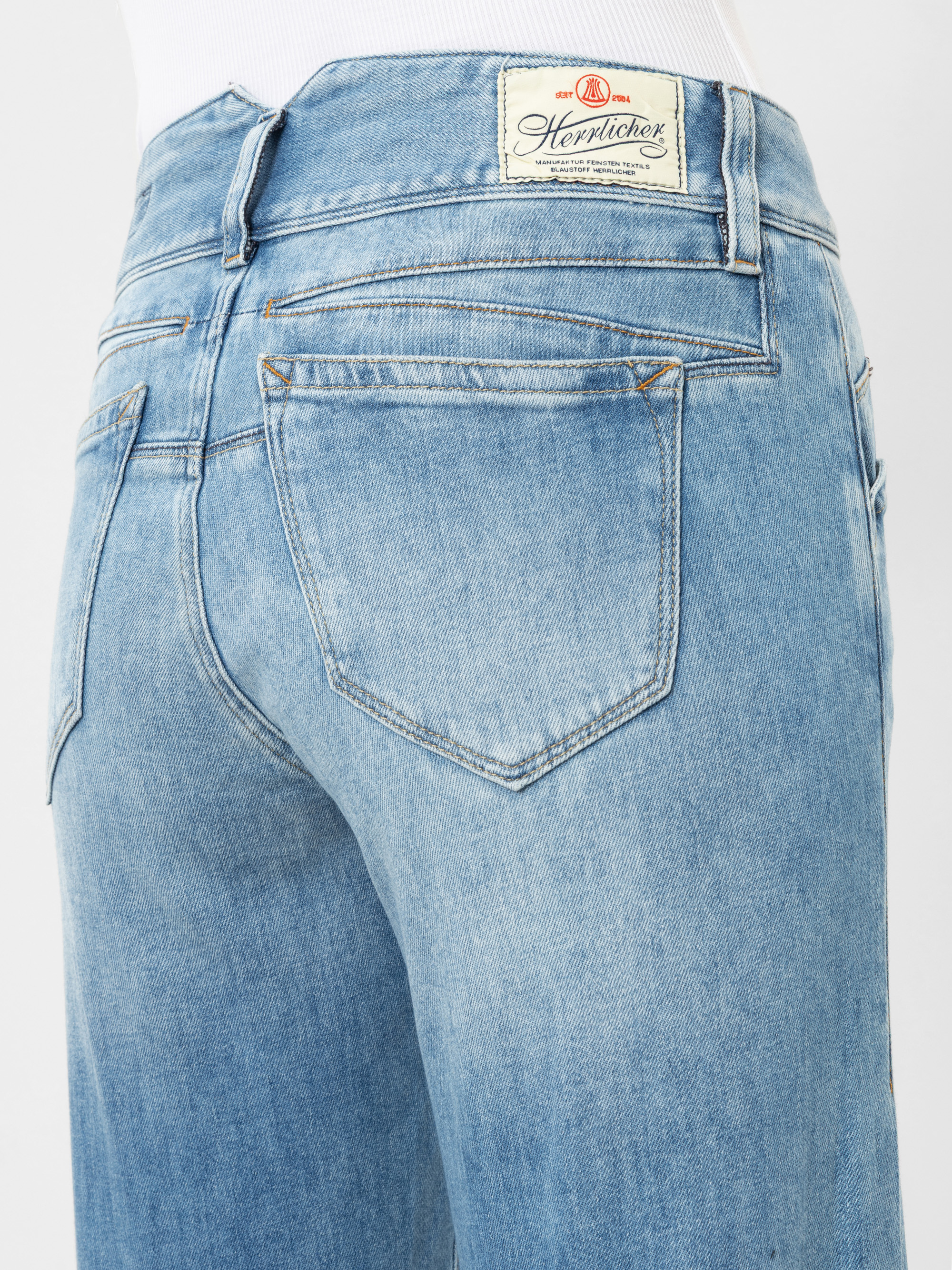 Nahaufnahme des Rückens einer Person, die eine hellblaue Jeans mit sichtbaren Gesäßtaschen und einem weißen Etikett mit der Aufschrift Herrlicher am Bund trägt. Die Person trägt außerdem ein weißes Oberteil.