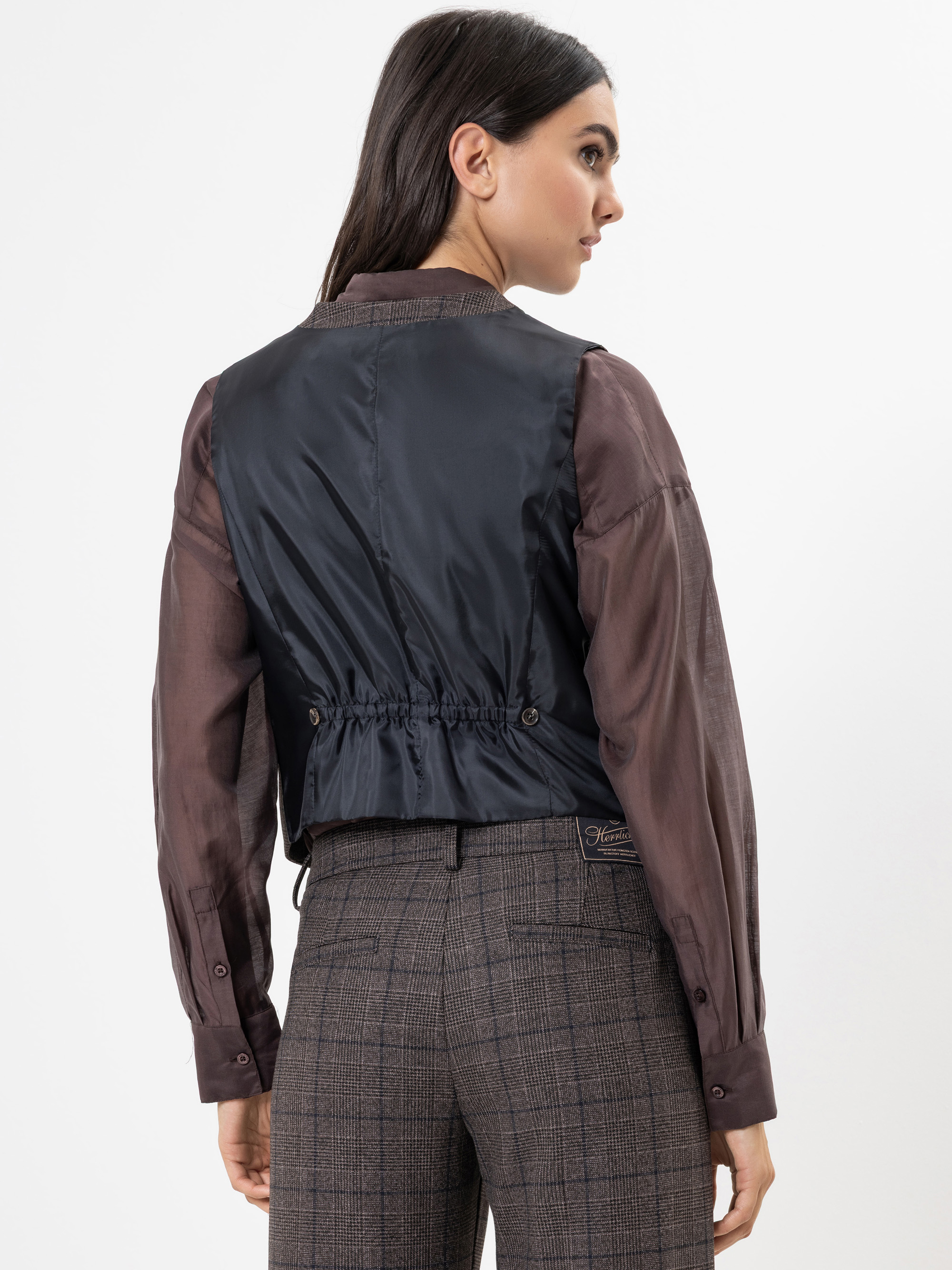 Eine Frau mit langen dunklen Haaren ist von hinten zu sehen. Sie trägt eine schwarze ärmellose Weste über einer durchsichtigen braunen langärmeligen Bluse und einer hochgeschnittenen karierten Hose. Das Outfit ist stilvoll und modern vor einem schlich