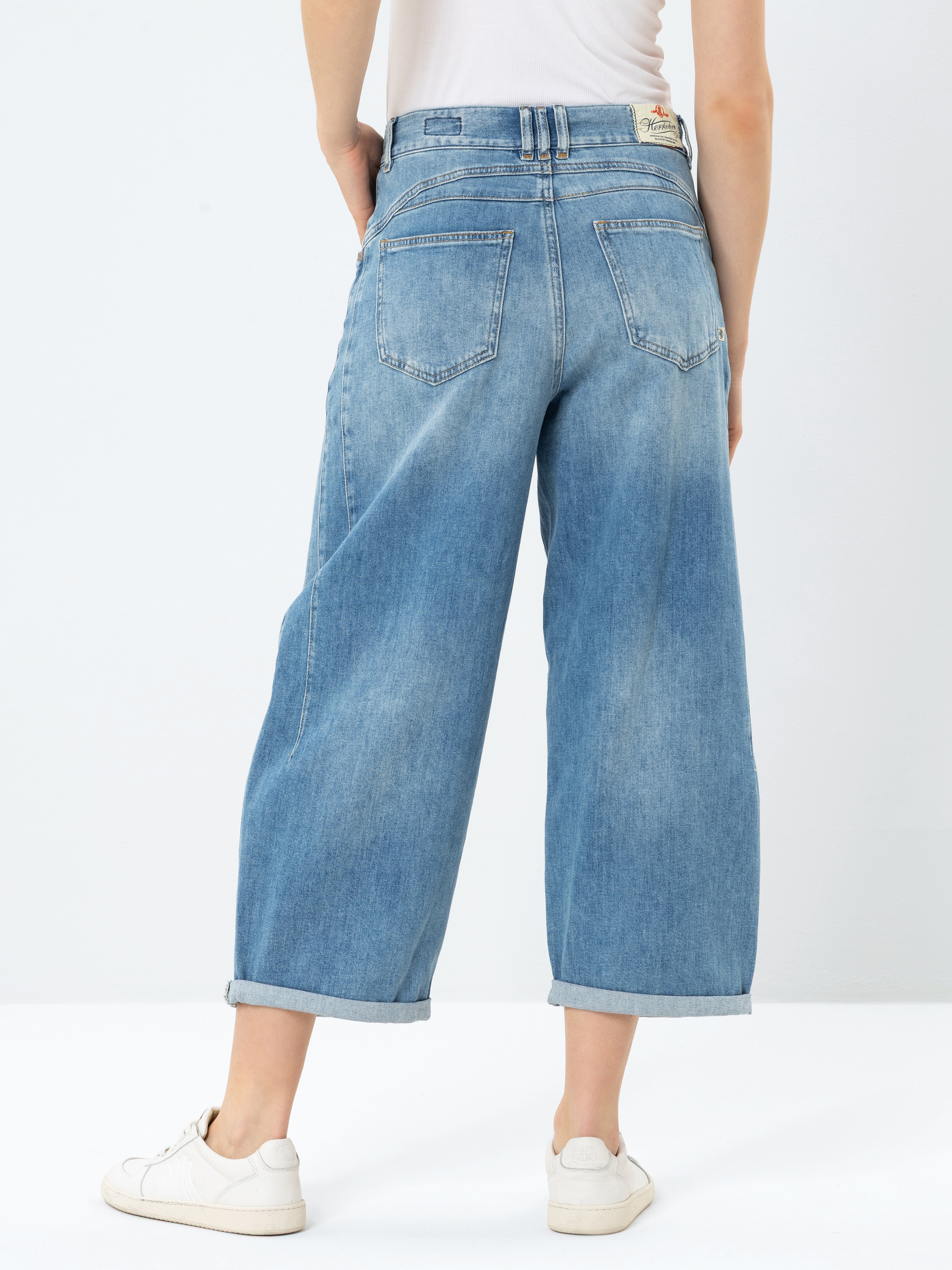 Eine Person, die hellblaue, weit geschnittene Jeans, ein weißes Oberteil und weiße Turnschuhe trägt, steht mit dem Rücken zur Kamera vor einem schlichten hellen Hintergrund.