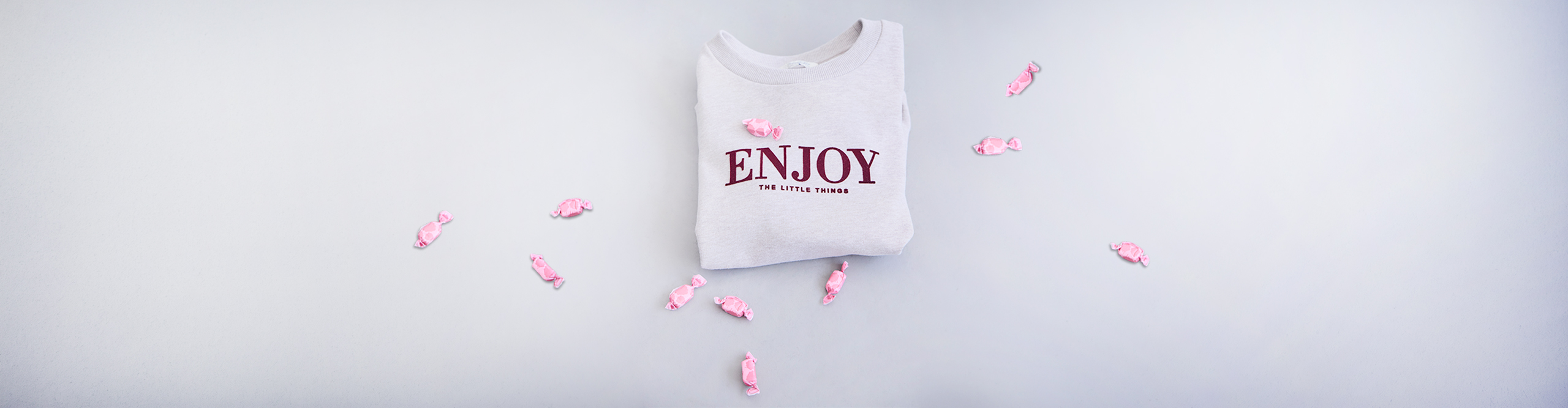 Ein hellgraues Sweatshirt mit dem Aufdruck ENJOY THE LITTLE THINGS, umrahmt von rosa verpackten Bonbons.