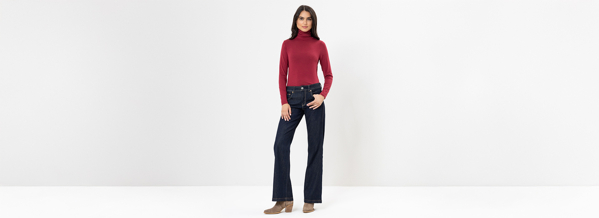 Eine Frau steht vor einem schlichten weißen Hintergrund und trägt einen roten Rollkragenpullover, dunkelblaue Jeans und hellbraune Ankle Boots. Sie hat glattes, schulterlanges dunkles Haar und eine entspannte, selbstbewusste Körperhaltung mit einer Han