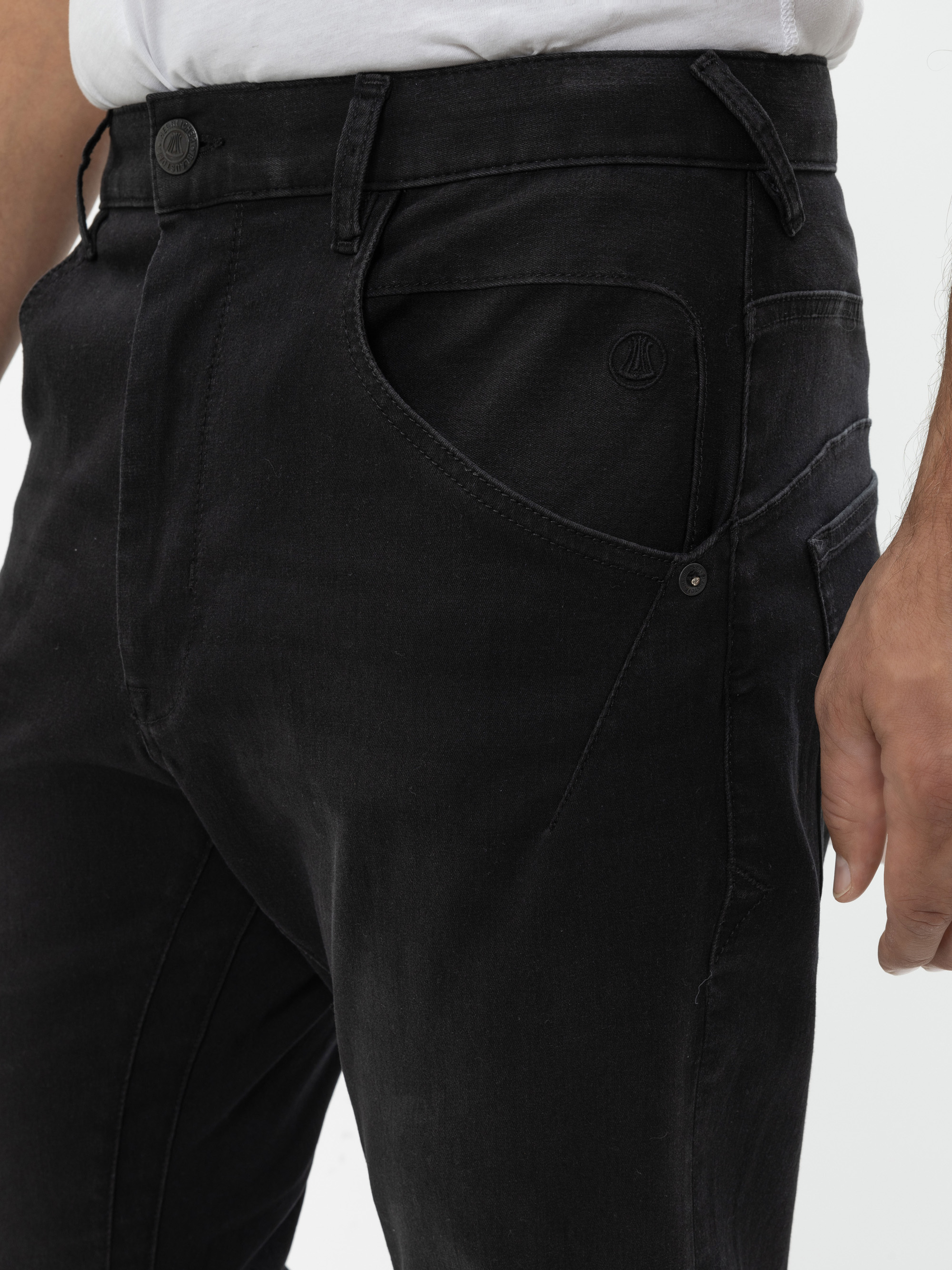 Eine Nahaufnahme einer Person, die eine schwarze Jeans und ein weißes T-Shirt trägt. Der Fokus liegt auf dem Bund und dem oberen Oberschenkelbereich, wobei die Nähte der Jeans, die Taschendetails und ein kleines Logo auf der Vordertasche hervorgehoben