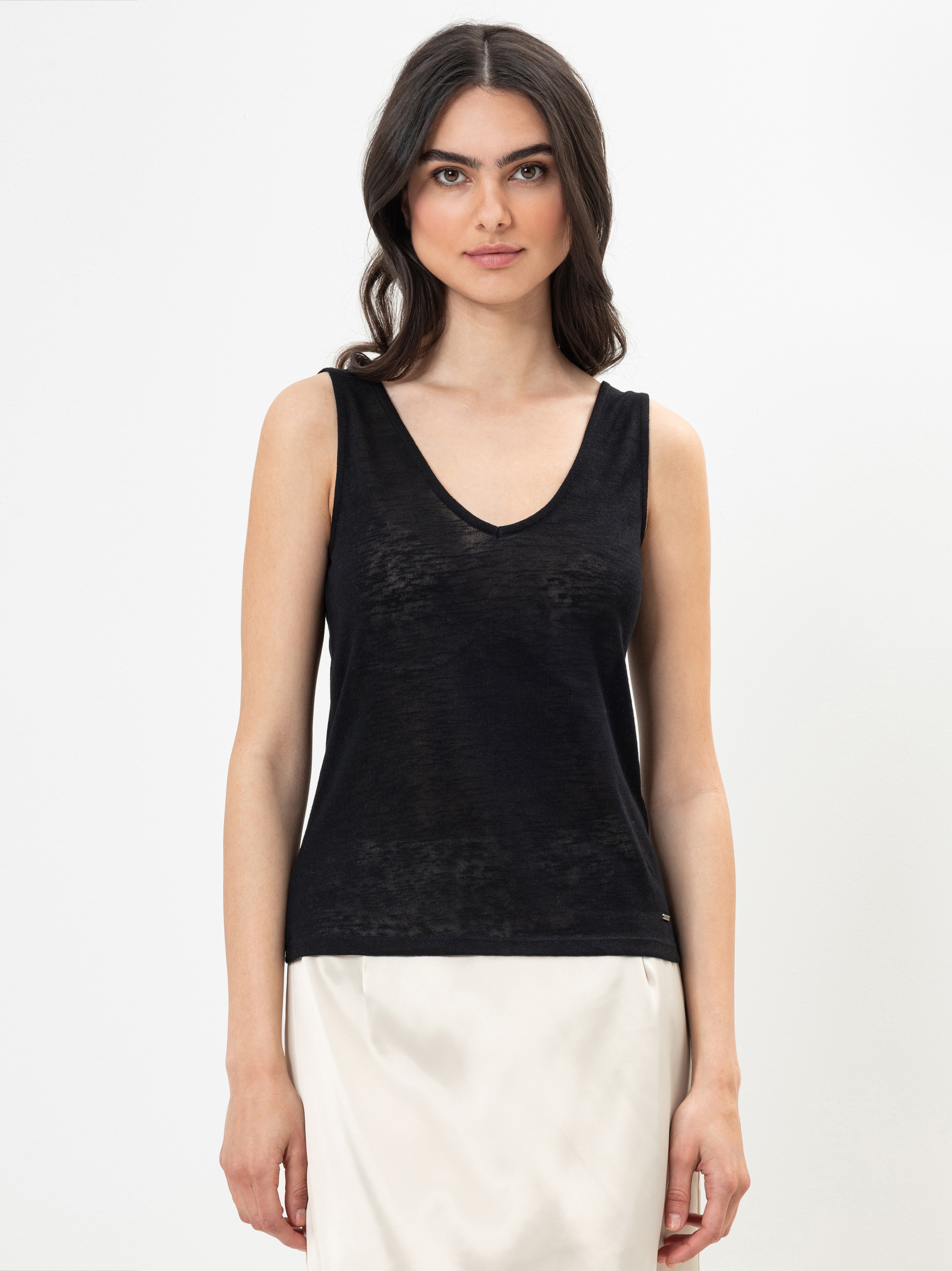 Eine Frau mit langen, dunklen Haaren trägt ein ärmelloses schwarzes Oberteil mit V-Ausschnitt und einen hellen Rock und steht vor einem schlichten weißen Hintergrund. Sie blickt nach vorne und hat einen neutralen Gesichtsausdruck.