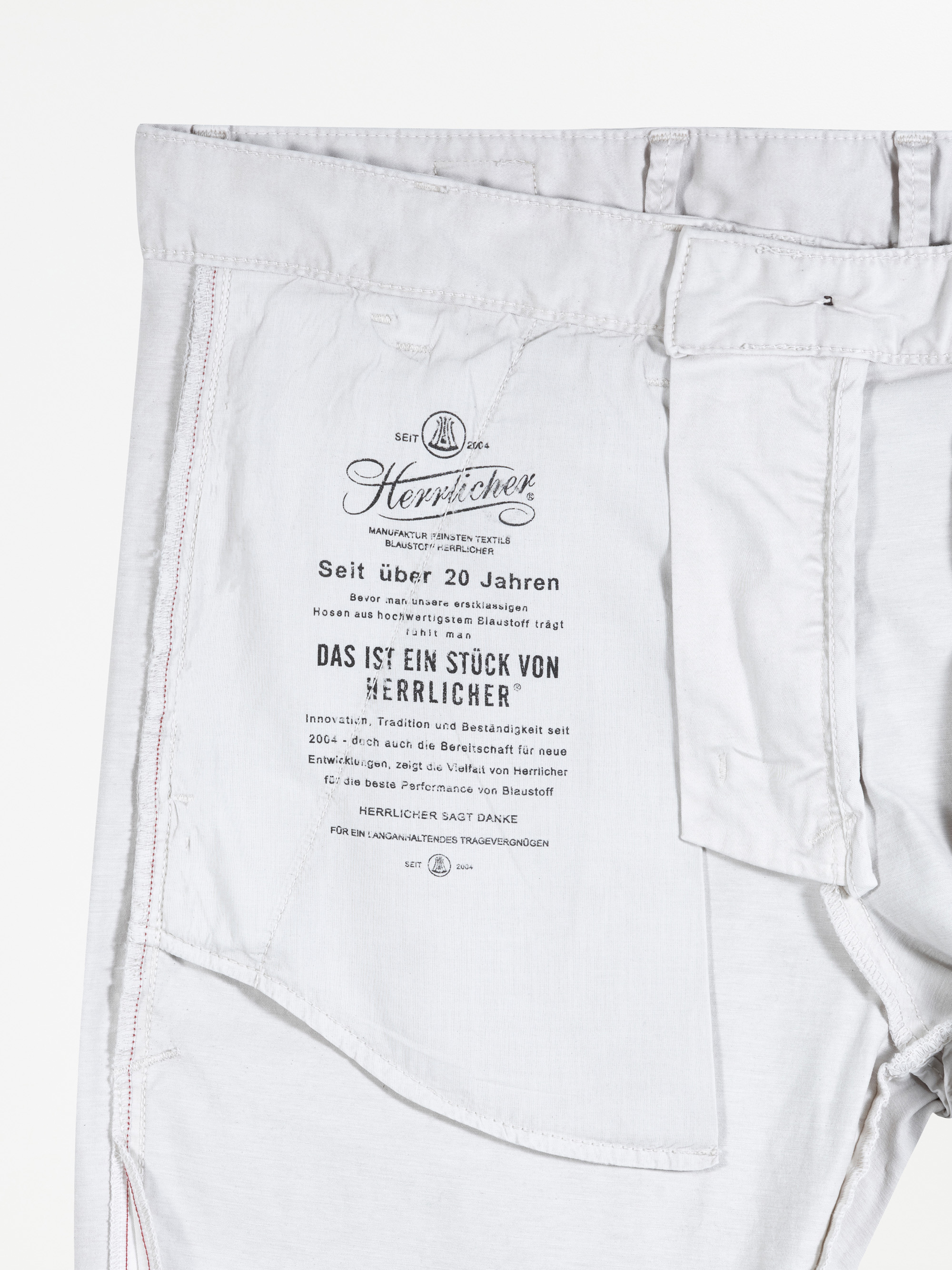 Die Innenseite der hellgrauen Hose zeigt ein gedrucktes Etikett mit dem Herrlicher-Markenlogo und einem deutschen Text, der die Geschichte der Marke und Produktinformationen beschreibt. Die Nähte und das Taschenfutter sind ebenfalls sichtbar.