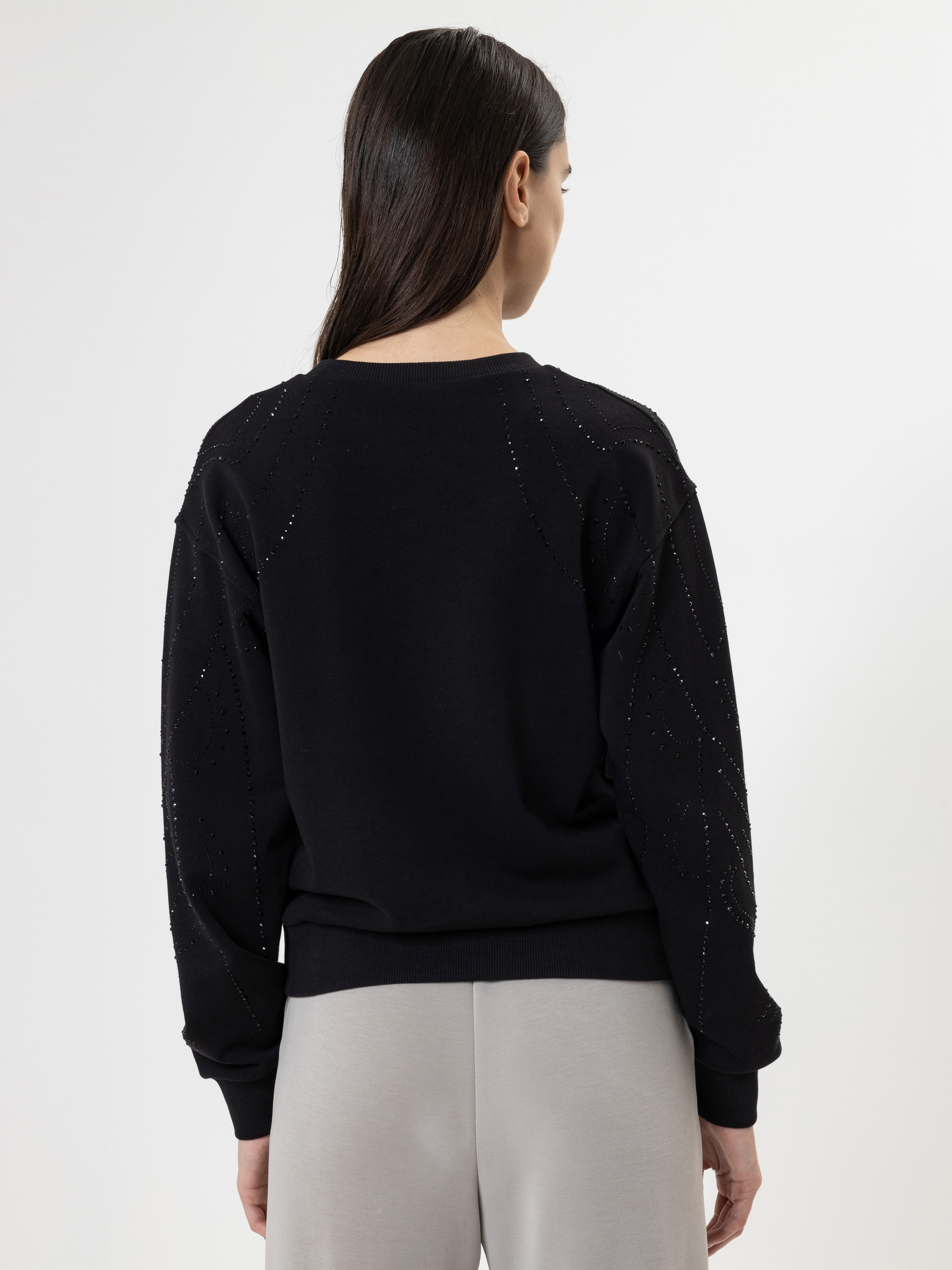 Eine Frau mit langen braunen Haaren, die einen schwarzen Pullover mit dezenten Verzierungen an den Ärmeln und eine hellgraue Hose trägt, steht mit dem Rücken zur Kamera vor einem schlichten weißen Hintergrund.
