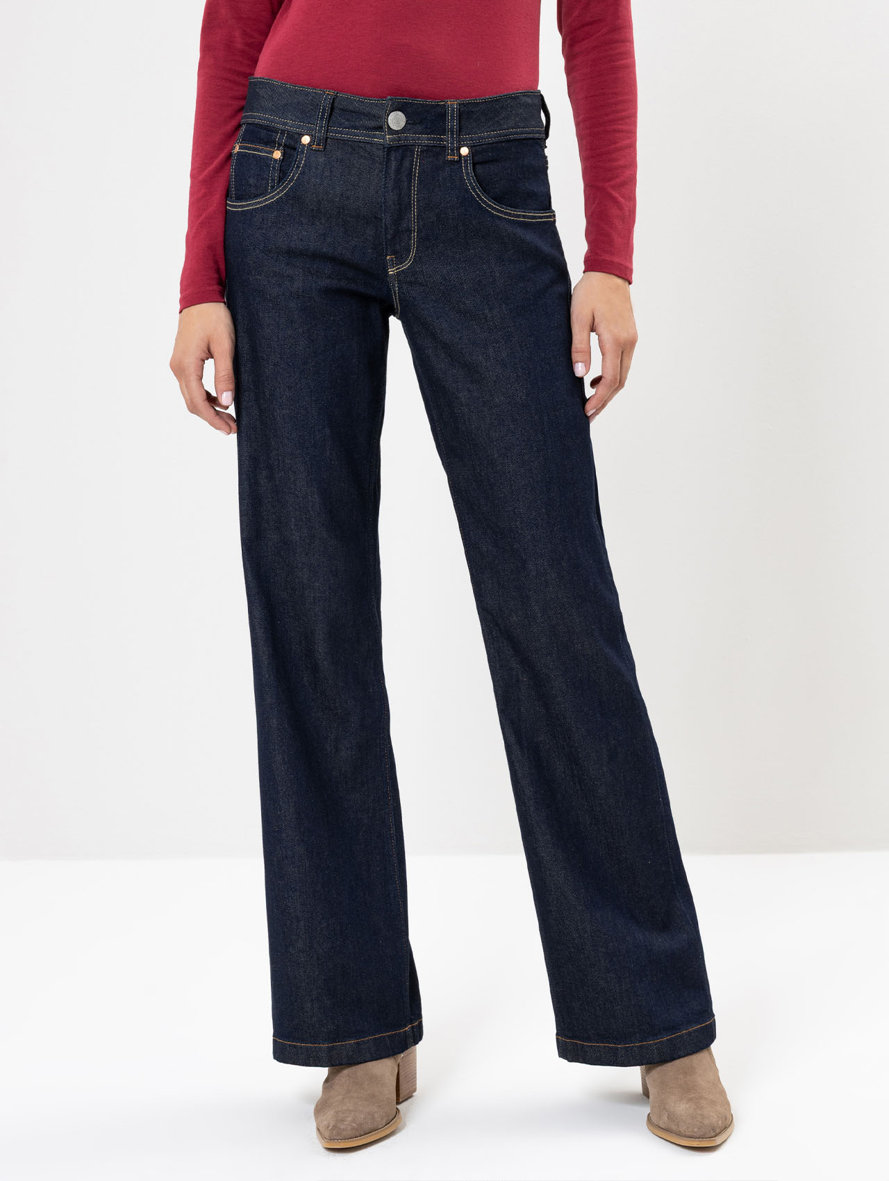 Person, die dunkelblaue Jeans mit weitem Bein, ein langärmeliges rotes Oberteil und beigefarbene Wildlederstiefel trägt und vor einem schlichten weißen Hintergrund steht. Der Kopf der Person ist auf dem Bild nicht zu sehen.