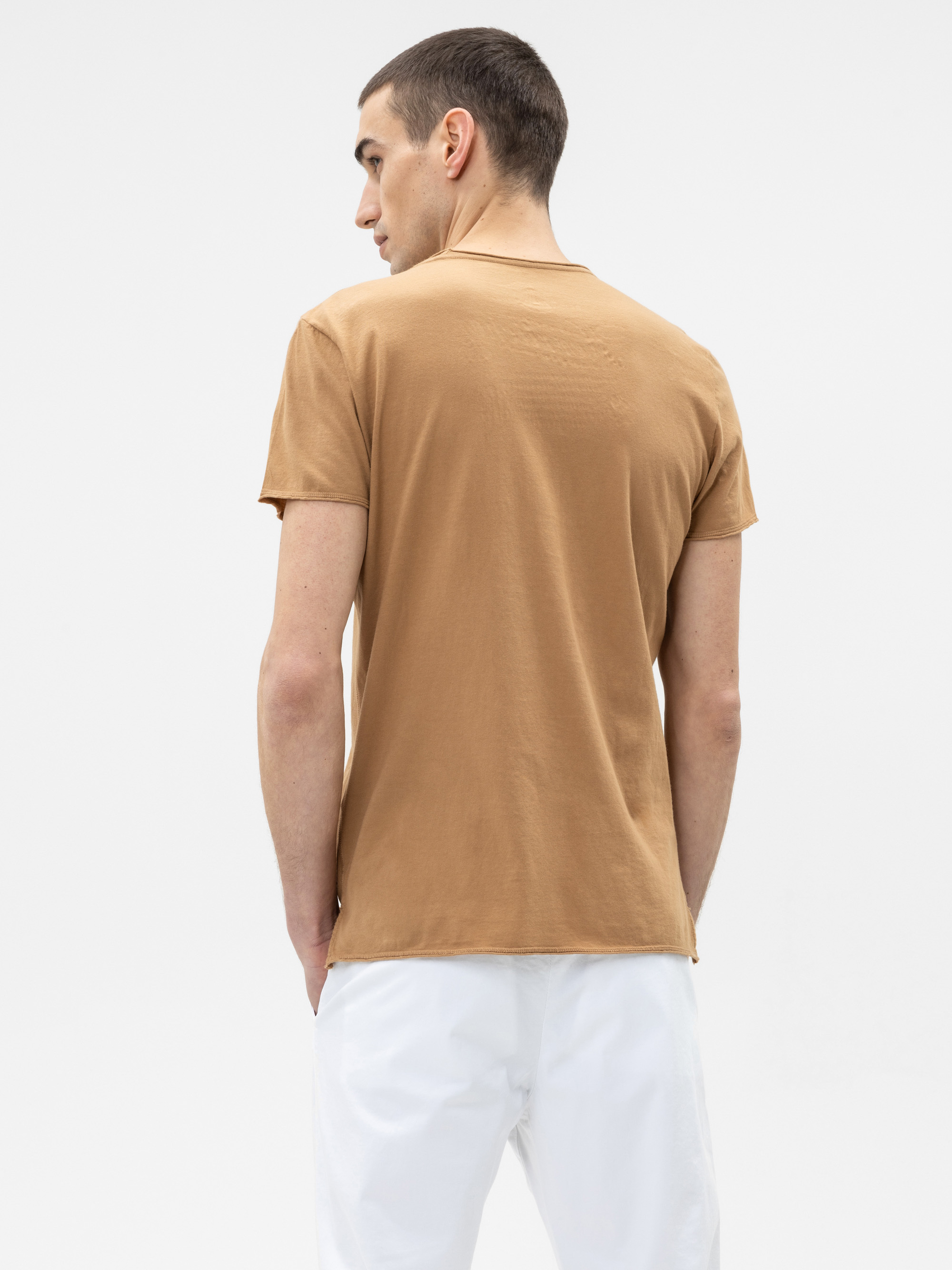 Ein Mann, der ein hellbraunes kurzärmeliges T-Shirt und eine weiße Hose trägt, steht mit dem Rücken zur Kamera vor einem schlichten weißen Hintergrund.