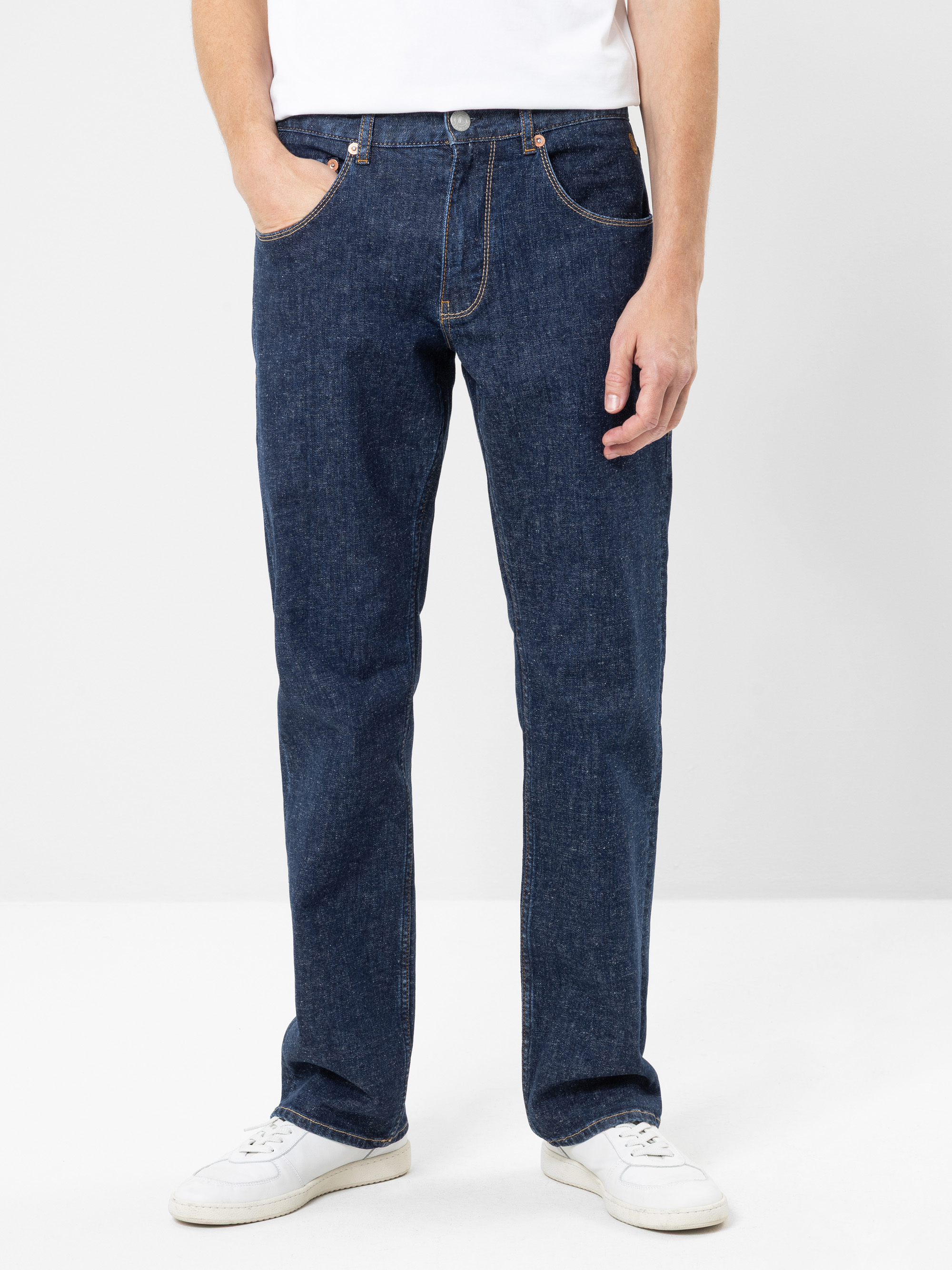 Eine Person, die eine dunkelblaue Jeans mit geradem Bein, ein weißes T-Shirt und weiße Turnschuhe trägt, steht vor einem schlichten hellen Hintergrund und hat eine Hand in der Hosentasche.