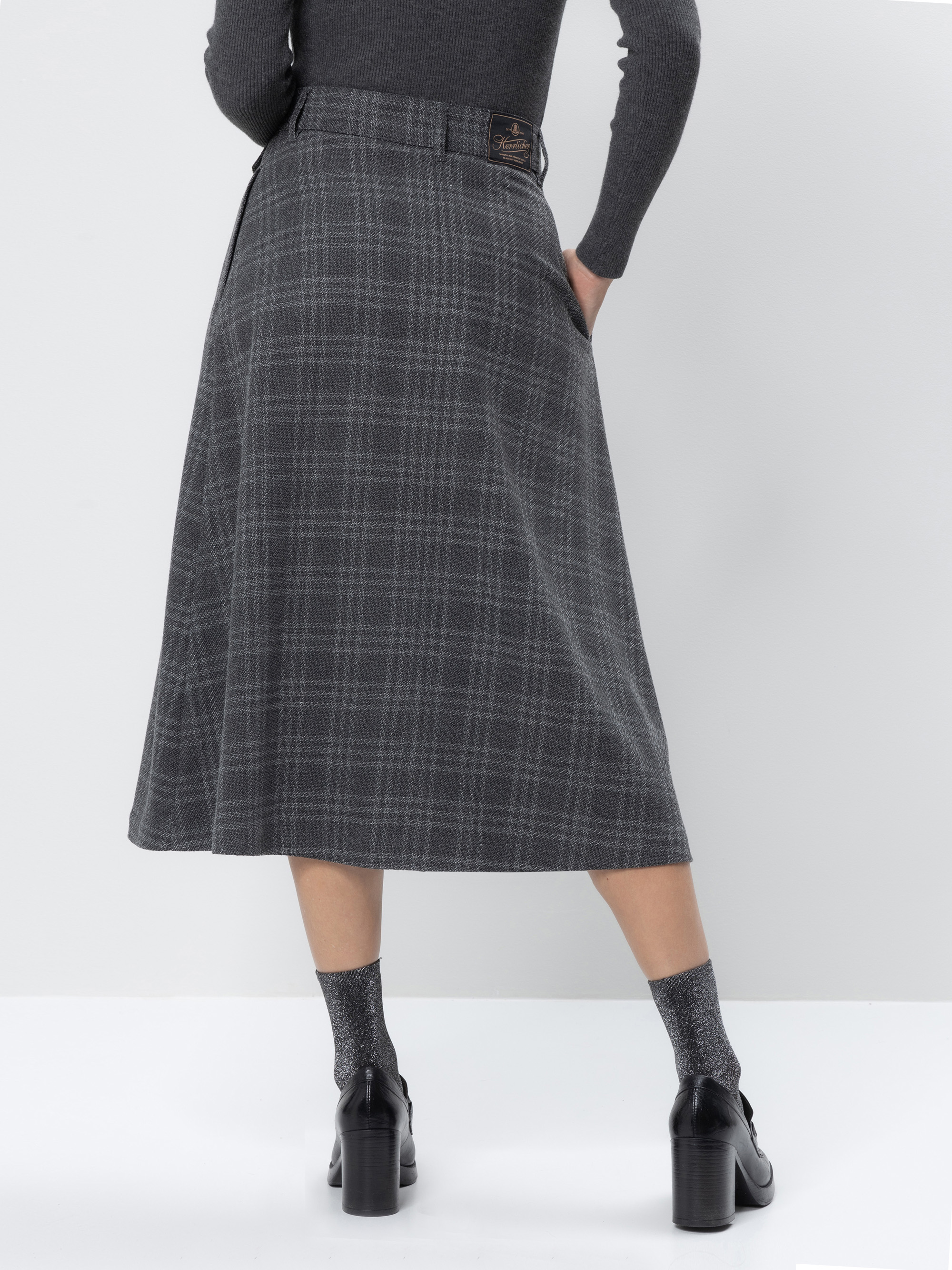 Eine Person, die einen grau karierten Midirock, graue Socken und schwarze hochhackige Schuhe trägt, steht vor einem hellen Hintergrund, von hinten gesehen.