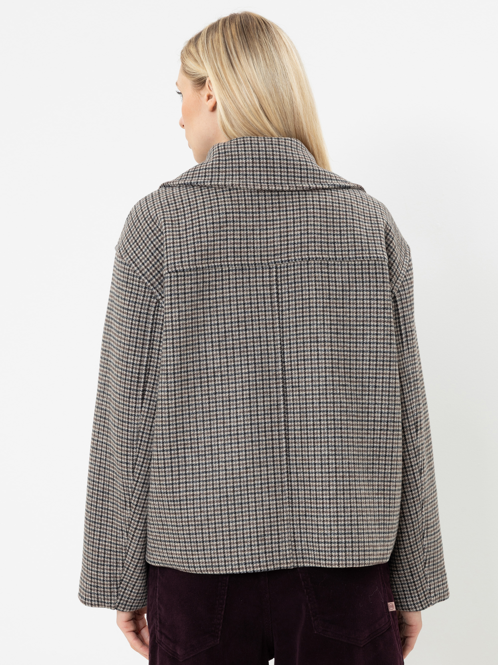 Eine Person mit langen blonden Haaren, von hinten gesehen, trägt eine locker sitzende, braun-grau karierte Jacke und eine dunkle Hose und steht vor einem schlichten weißen Hintergrund.