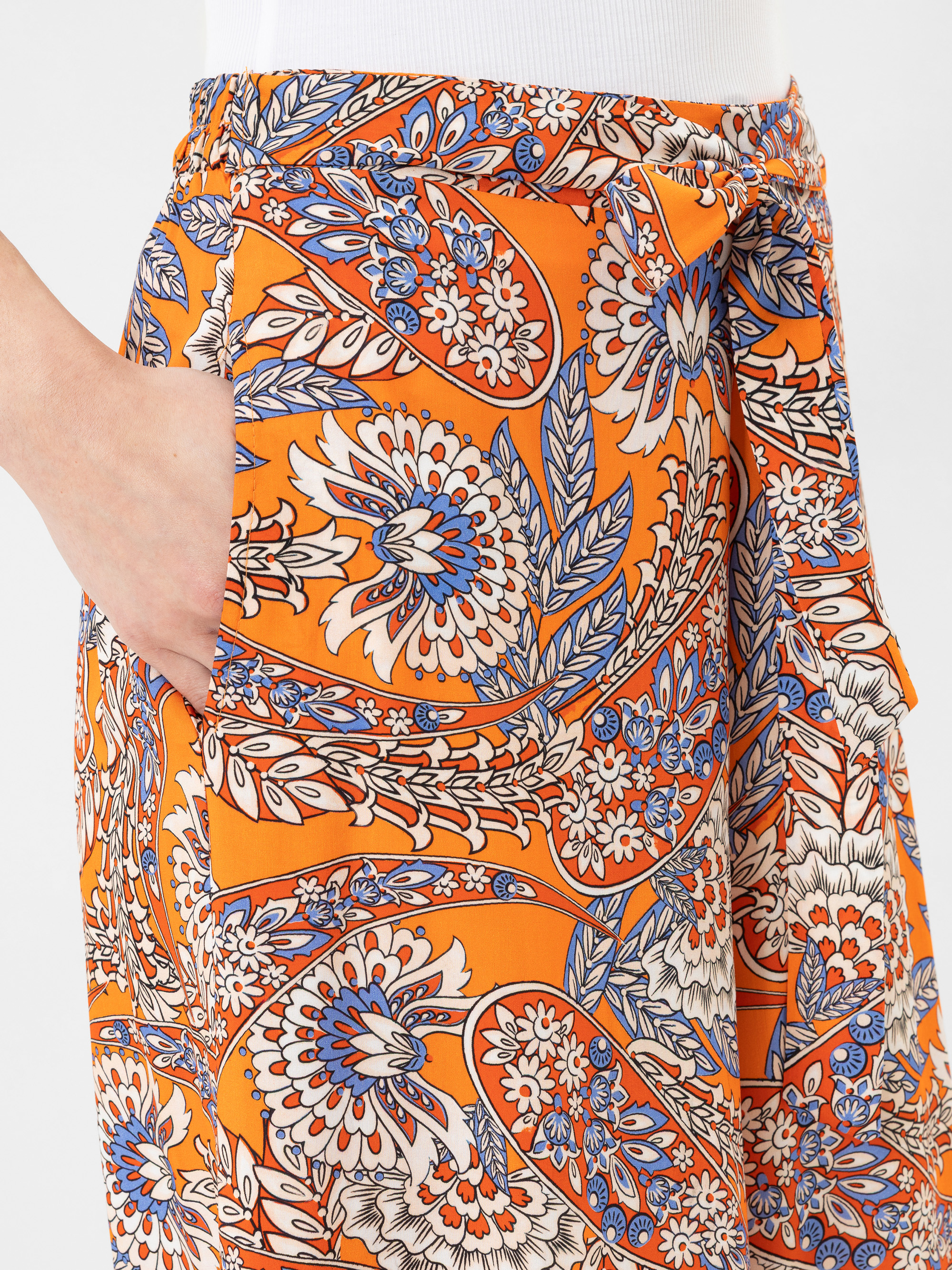 Eine Person trägt eine leuchtend orangefarbene Hose mit einem Blumen- und Blattmuster in Blau, Weiß und Rot und hat eine Hand in der Tasche. Die Hose hat eine hohe Taille und wird in der Taille mit einem passenden Stoffgürtel gebunden.