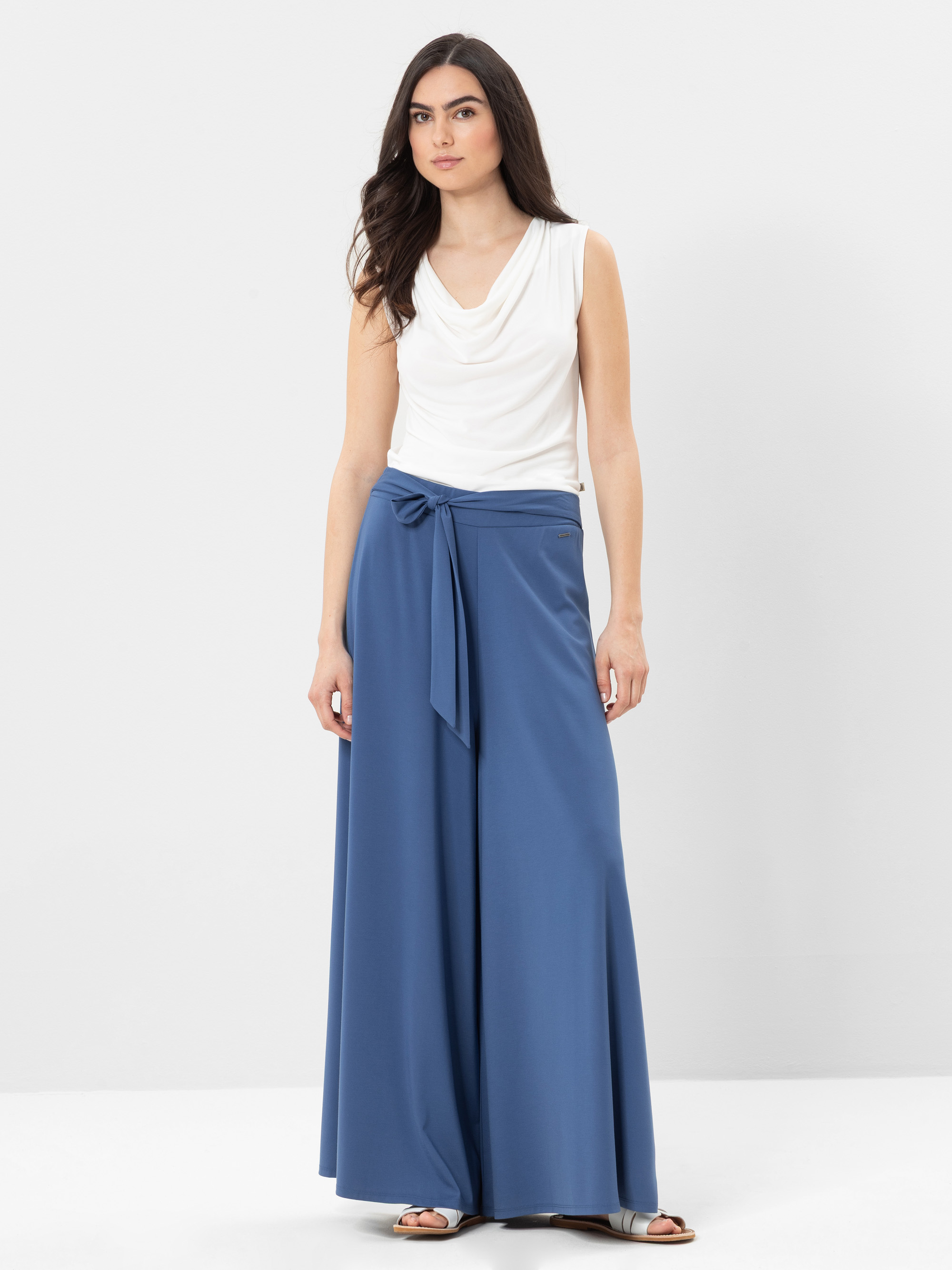 Eine Frau steht vor einem einfarbig weißen Hintergrund und trägt ein ärmelloses weißes Oberteil mit Kuhkragen und eine blaue Hose mit hoher Taille und weitem Bein, die mit einer Krawatte gebunden ist. Sie hat langes dunkles Haar und trägt Sandalen.